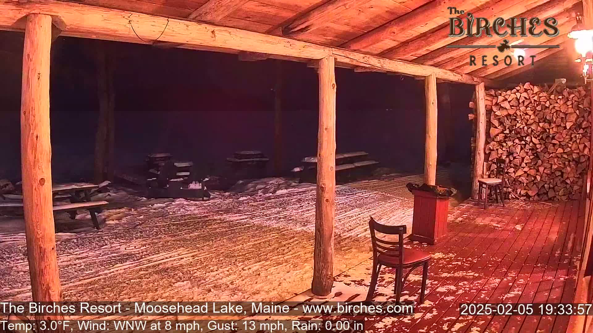 A snow-covered outdoor patio with picnic tables and chairs under a wooden covered structure at night.