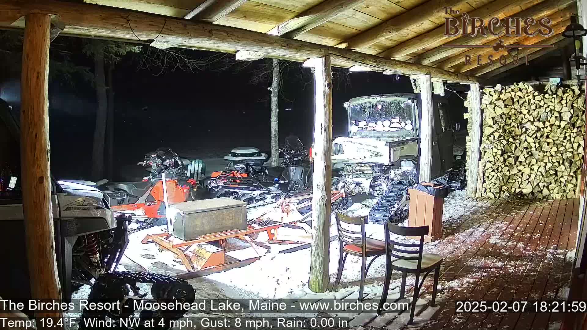 Under a covered wooden porch at night in snowy conditions, several snowmobiles and other motorized vehicles are parked, with a large stack of firewood visible to the right.