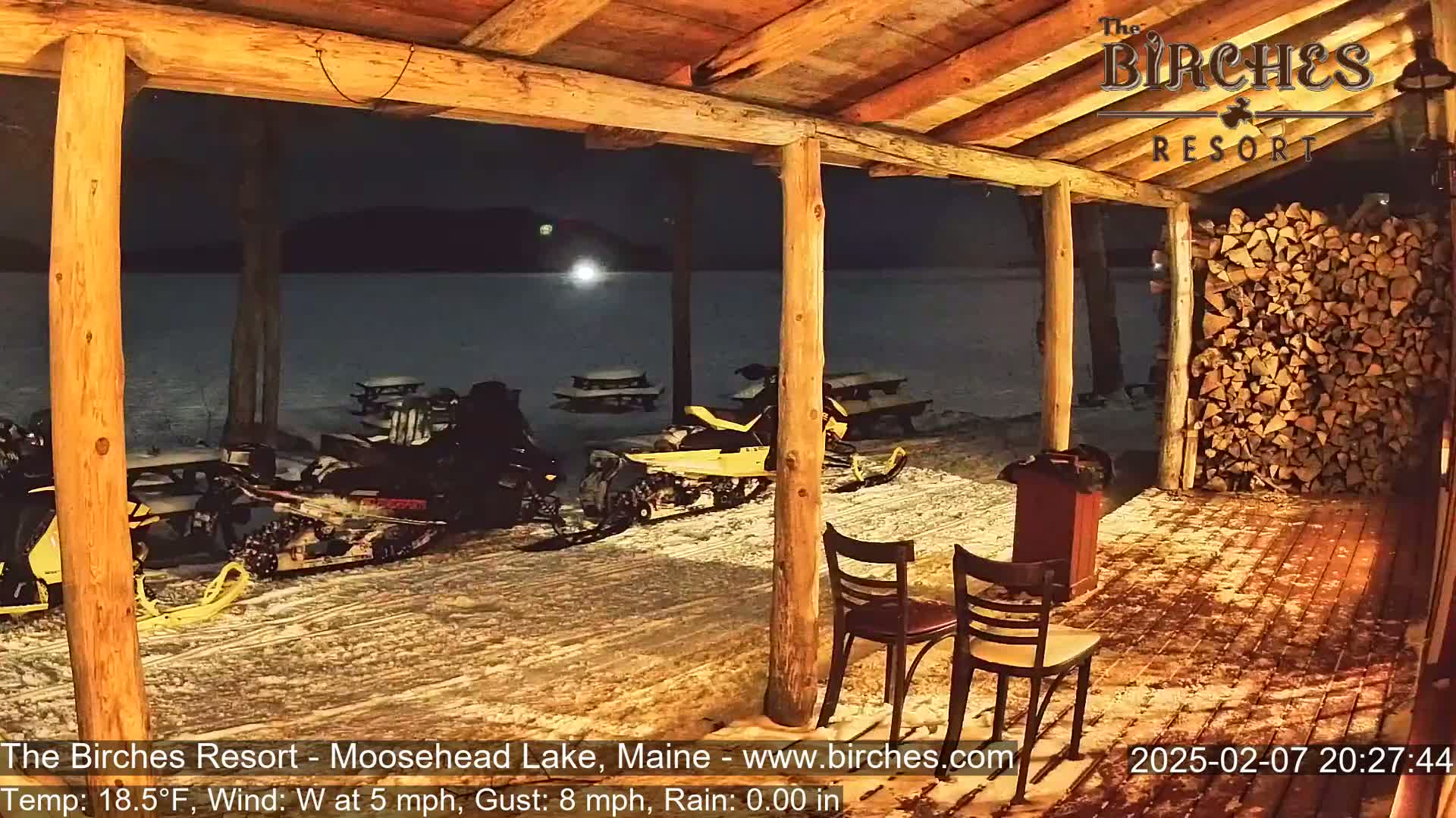 Snowmobiles are parked outside a wooden porch at night in snowy conditions with a west wind.