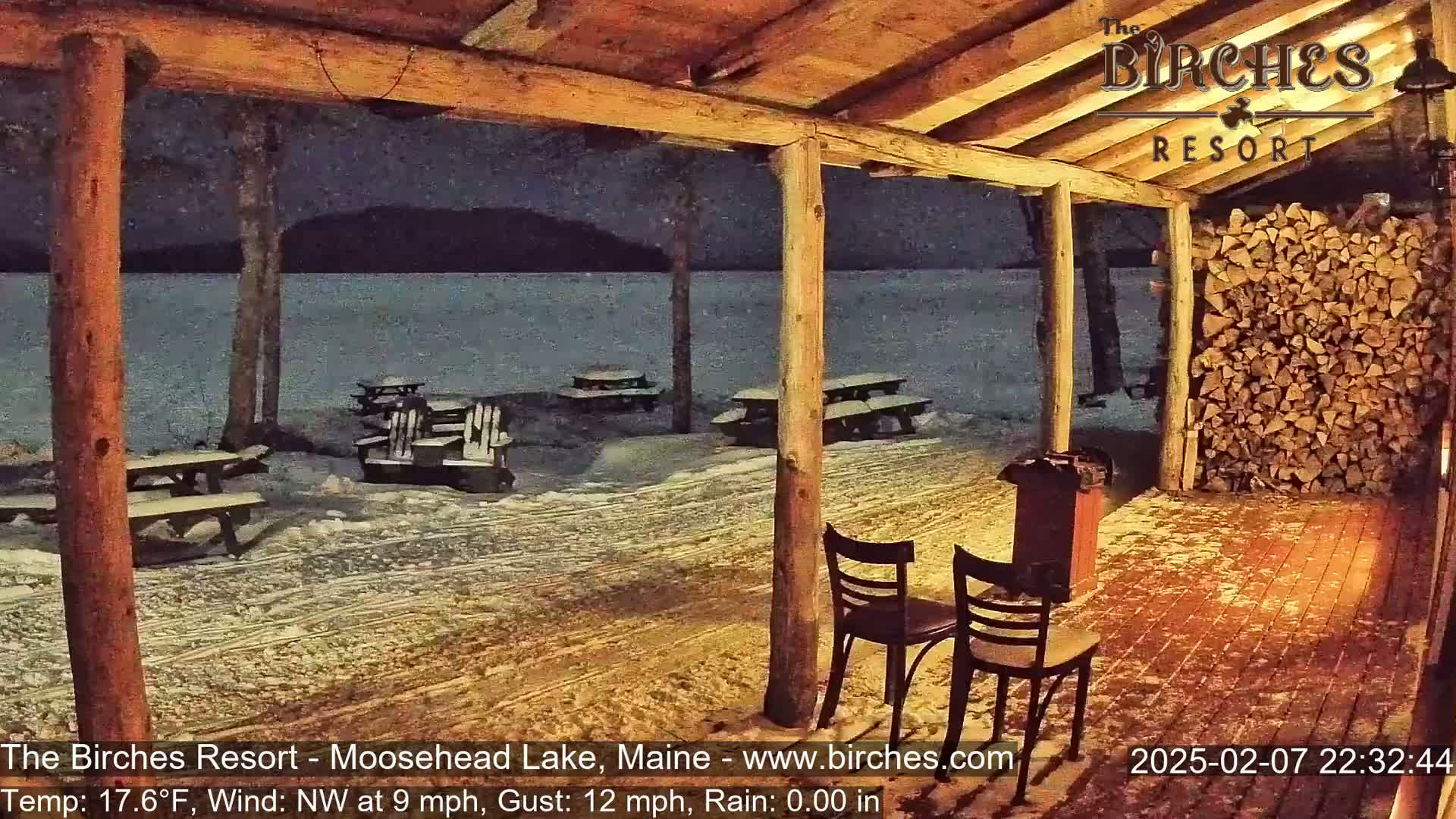 A snow-covered outdoor patio with picnic tables and chairs overlooks a dark lake under a nighttime sky.
