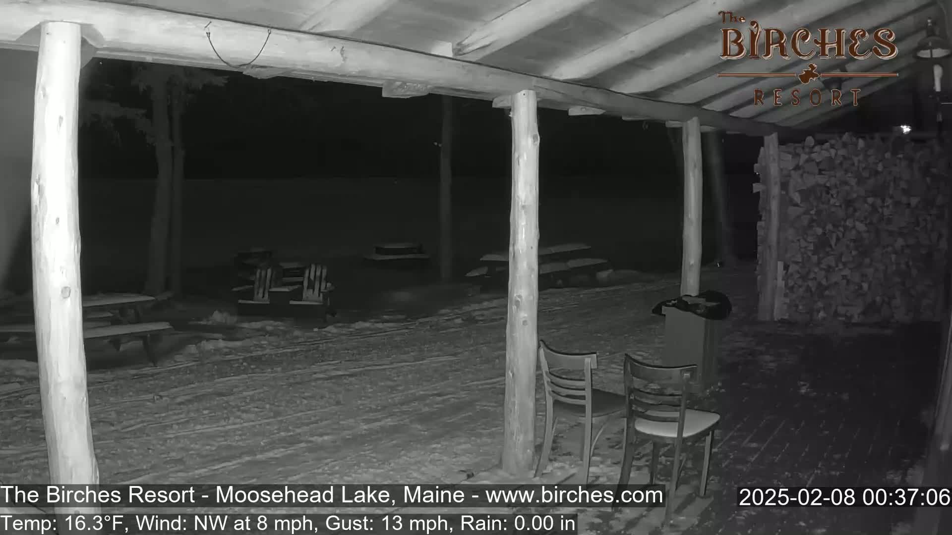 A snow-covered outdoor patio area with picnic tables and chairs is seen at night under a covered porch during a light Northwest wind and rain.