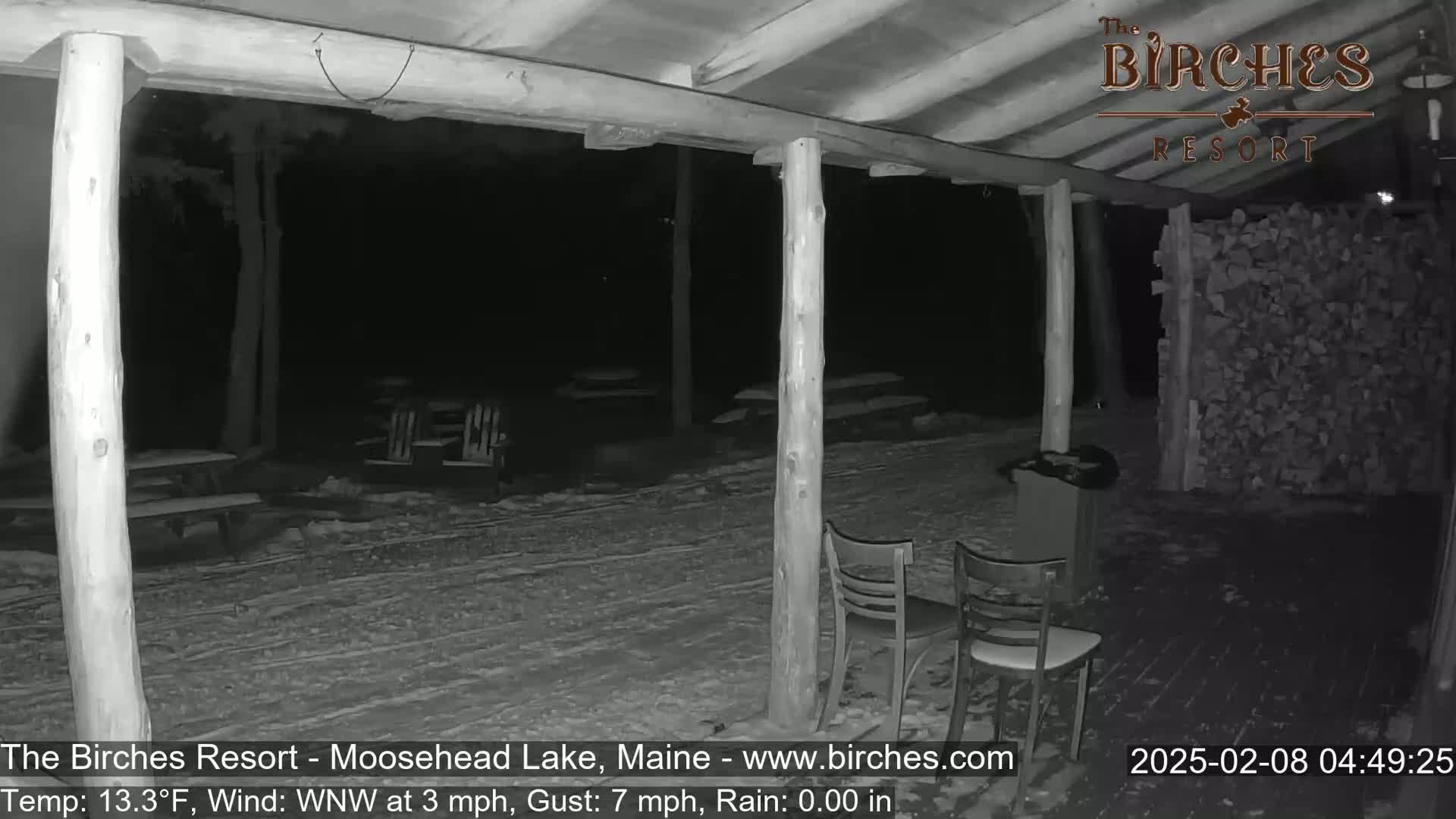 A snow-covered outdoor patio at night, partially sheltered by a wooden structure, with two empty chairs and a woodpile visible.