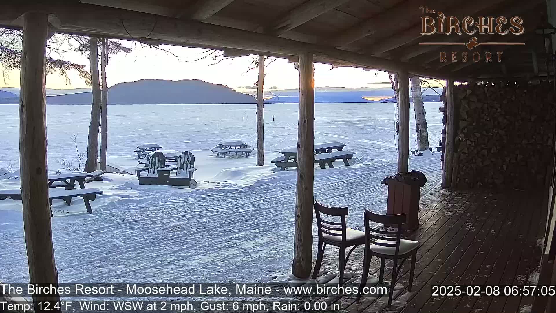 A snow-covered lakeside patio with picnic tables and chairs is viewed from a wooden porch, under a twilight sky.