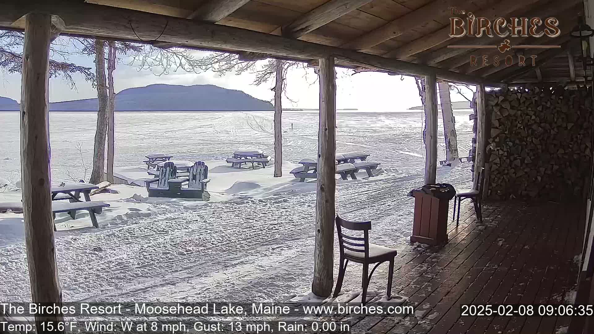 A snow-covered lakeside patio with picnic tables and chairs is viewed from a wooden porch on a partly sunny, cold, and windy day.