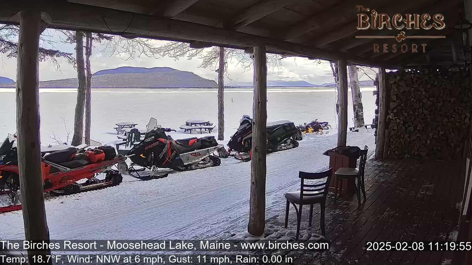 Several snowmobiles are parked outside a snow-covered lakeside porch with a view of mountains under a partly cloudy sky.