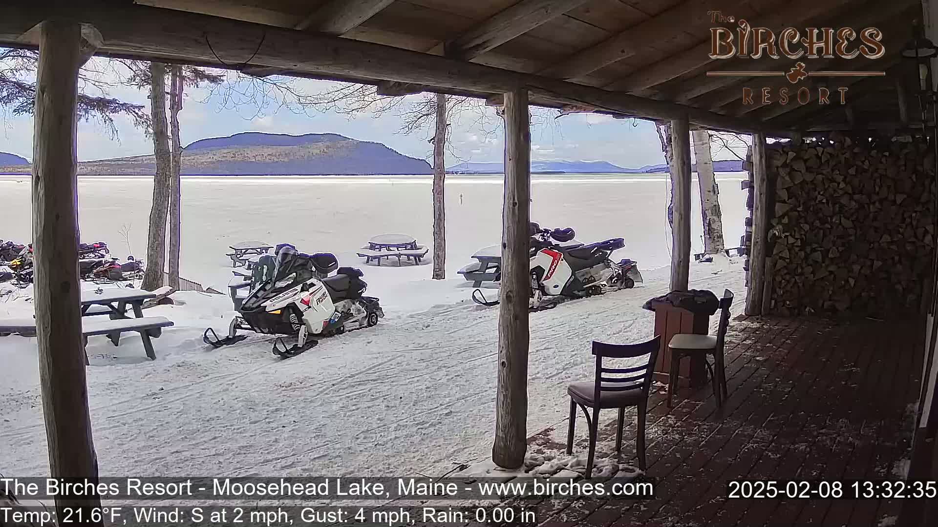 A snow-covered lake and mountains are viewed from a covered porch with snowmobiles parked nearby on a partly sunny day.