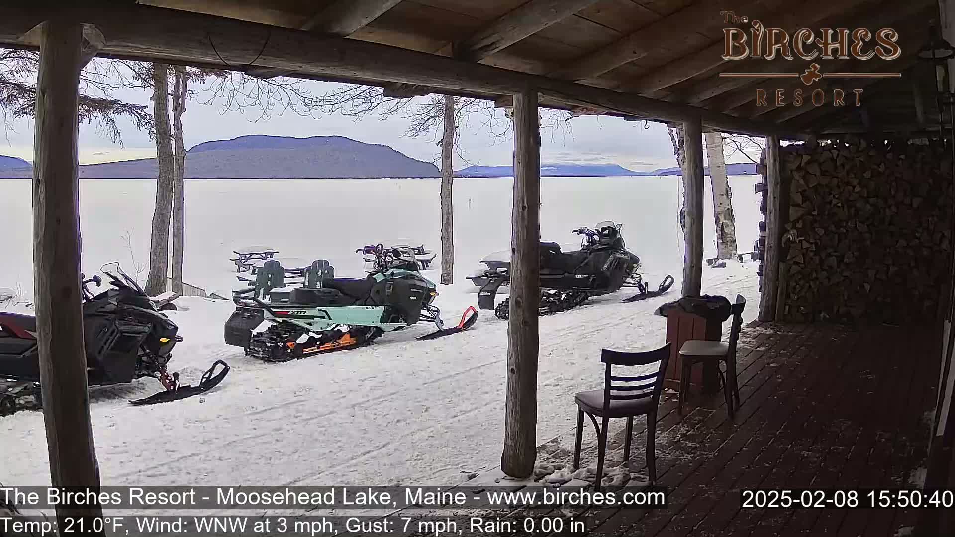 From a porch overlooking a snow-covered lake and distant mountains, several snowmobiles are parked in the snow.