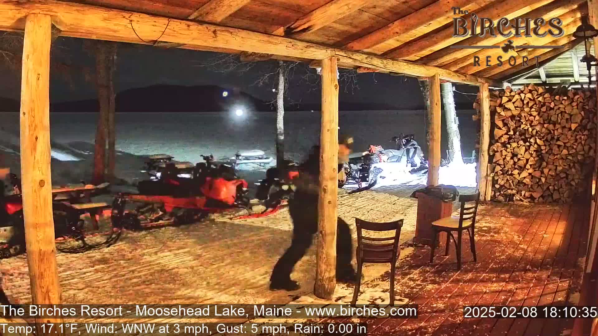 A person walks near snowmobiles parked outside a wooden porch at night, under a clear sky.