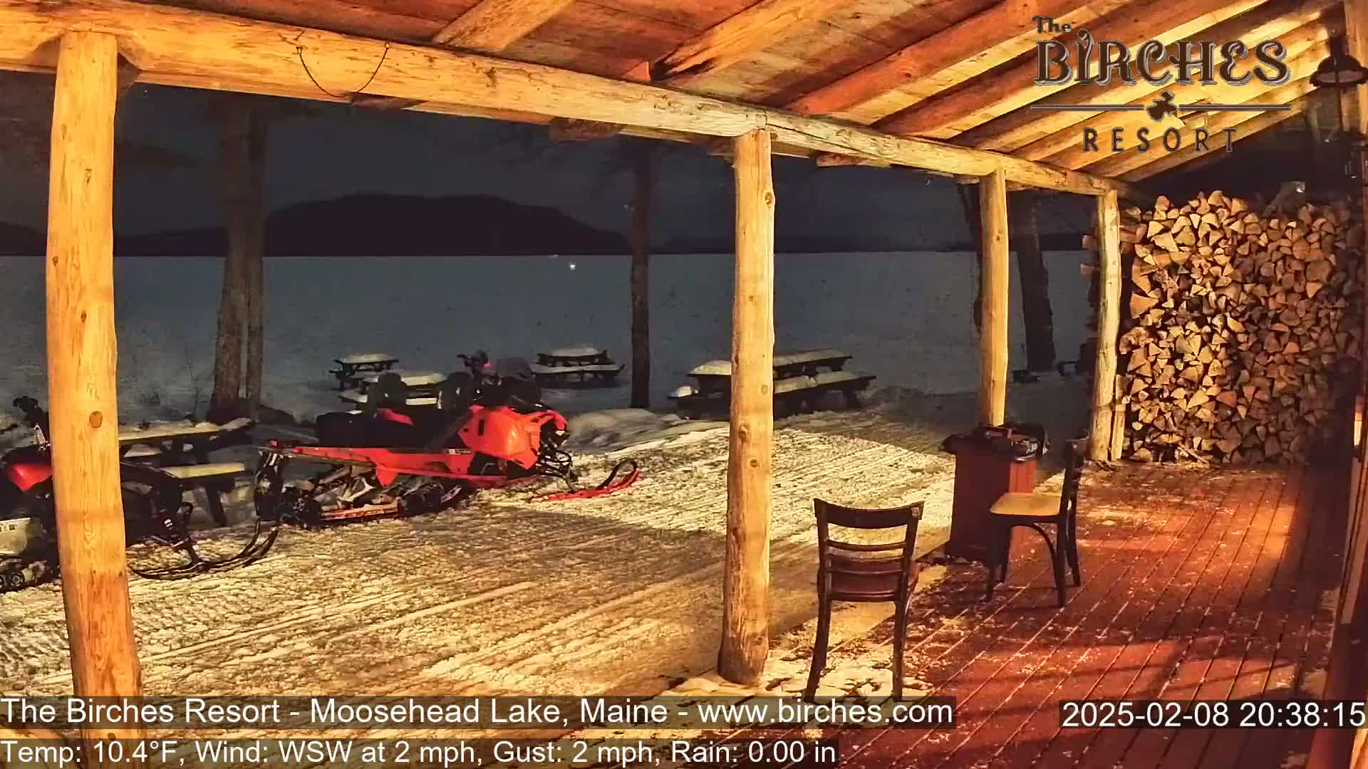 A snow-covered outdoor porch with snowmobiles parked nearby, viewed from inside a wooden structure at night.