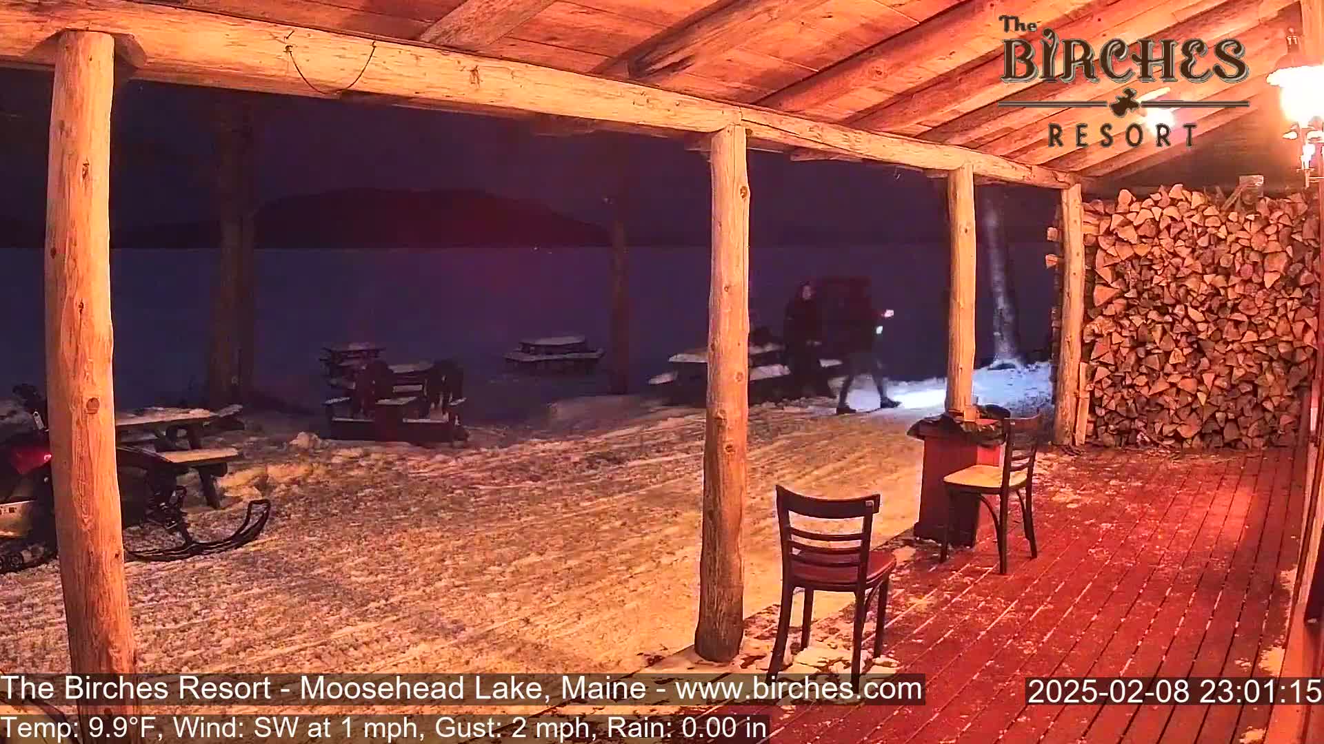 A snow-covered outdoor patio with wooden posts and picnic tables is illuminated at night, near a lake, with a few people walking by and a woodpile visible.
