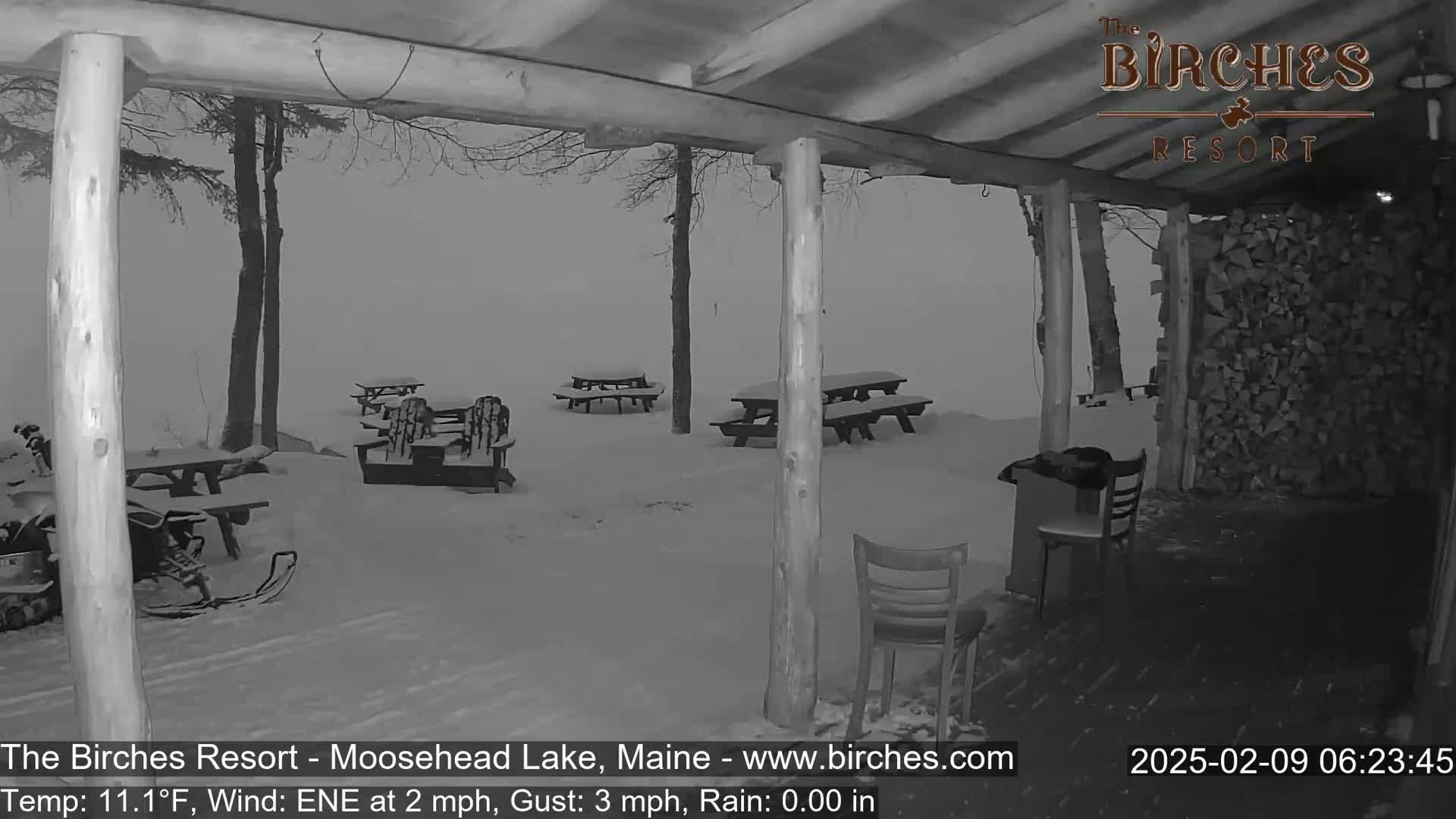 A snow-covered outdoor area with picnic tables and chairs is viewed from a covered porch during a rainy, windy day.