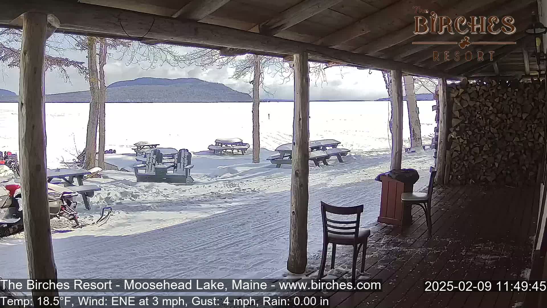 A snow-covered lakeside patio with picnic tables is viewed from a wooden porch on a partly cloudy day with a light wind and a temperature of 18.5°F.