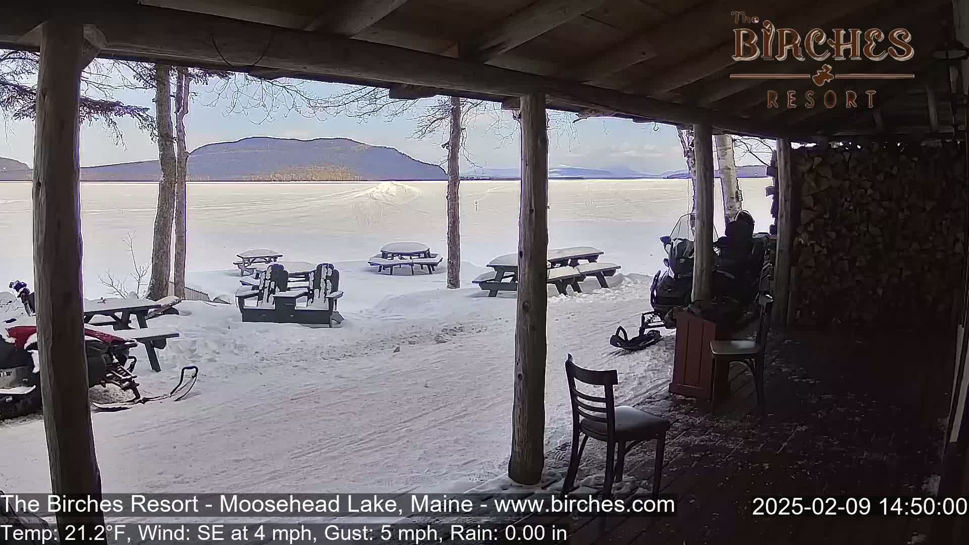 From a snow-covered porch overlooking a frozen lake and distant mountains, several snow-covered picnic tables and a snowmobile are visible under a clear, sunny sky.