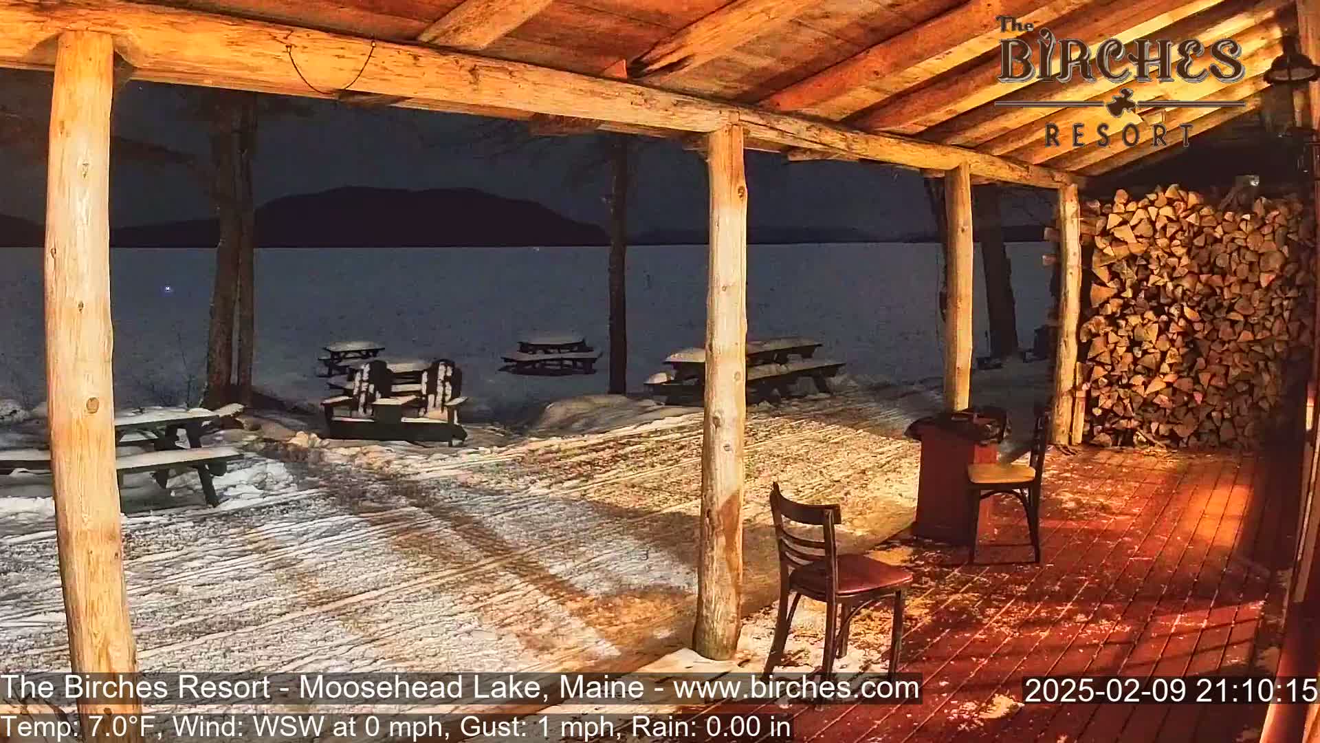 A snow-covered outdoor patio with picnic tables and chairs under a wooden structure overlooks a dark lake at night.