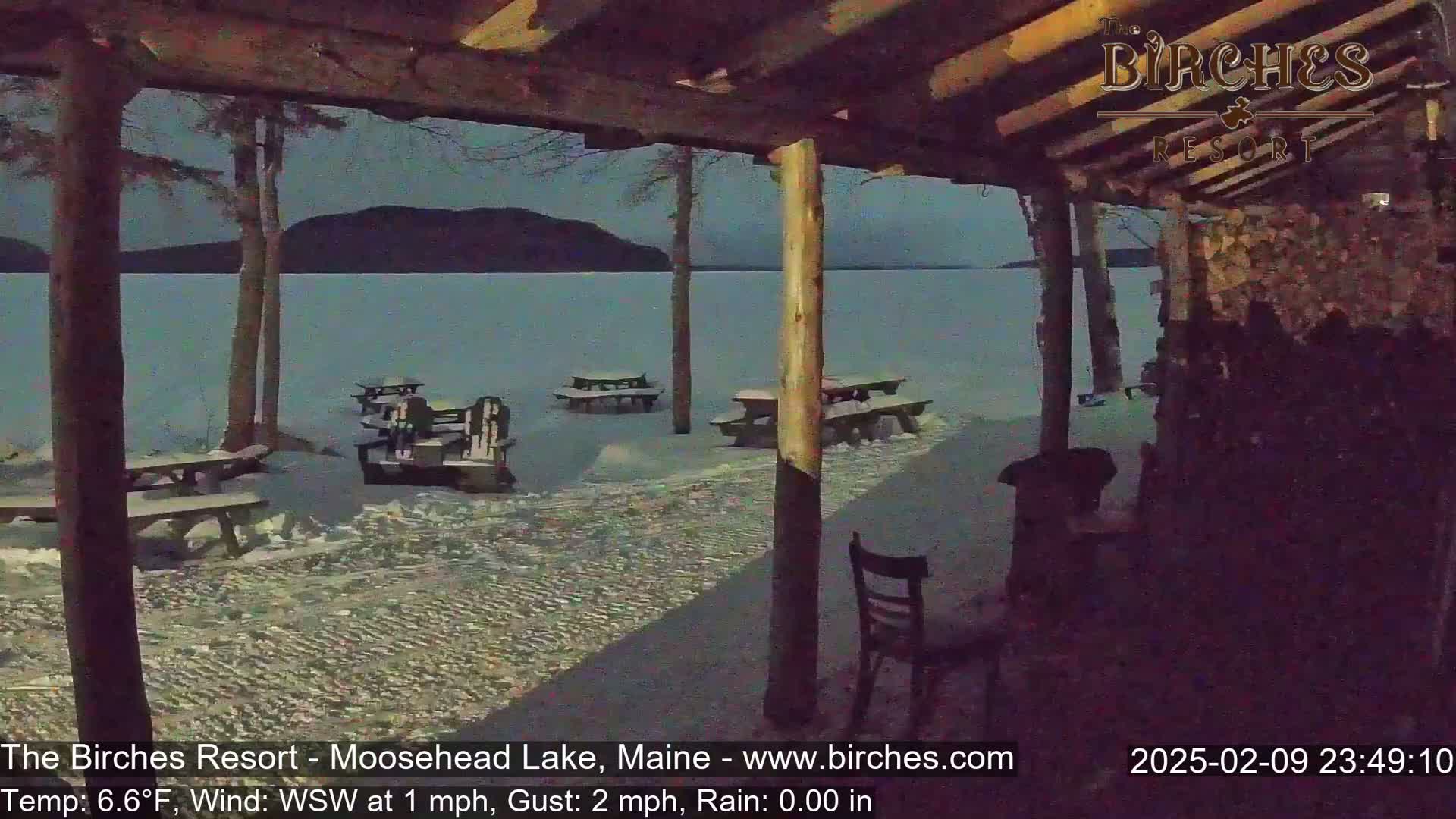 A snow-covered lakeside scene is viewed from under a covered porch at night, showing several empty picnic tables and chairs.