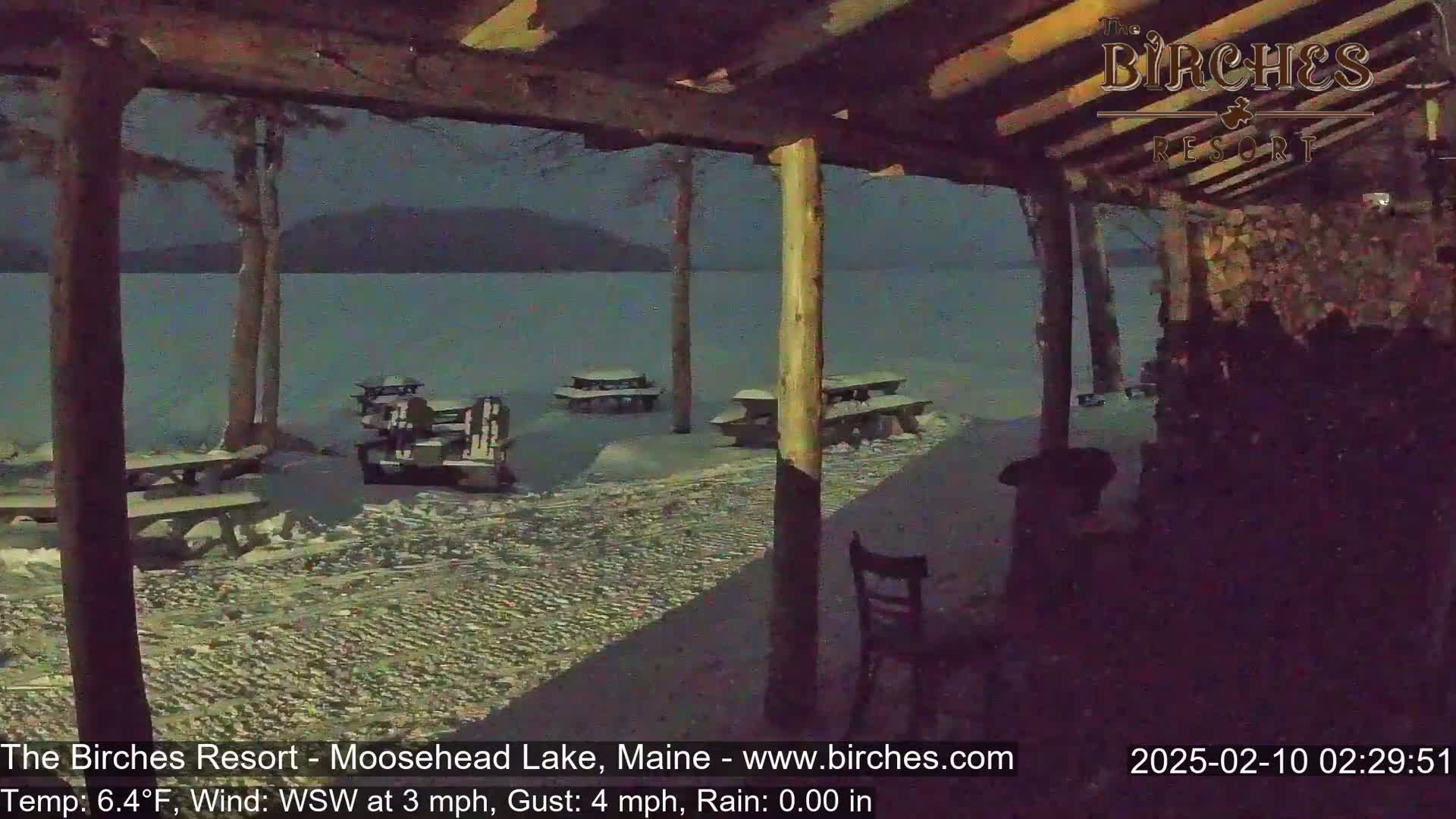 A snow-covered lakeside patio with picnic tables and chairs is seen at night under a covered wooden structure, during rainy conditions.