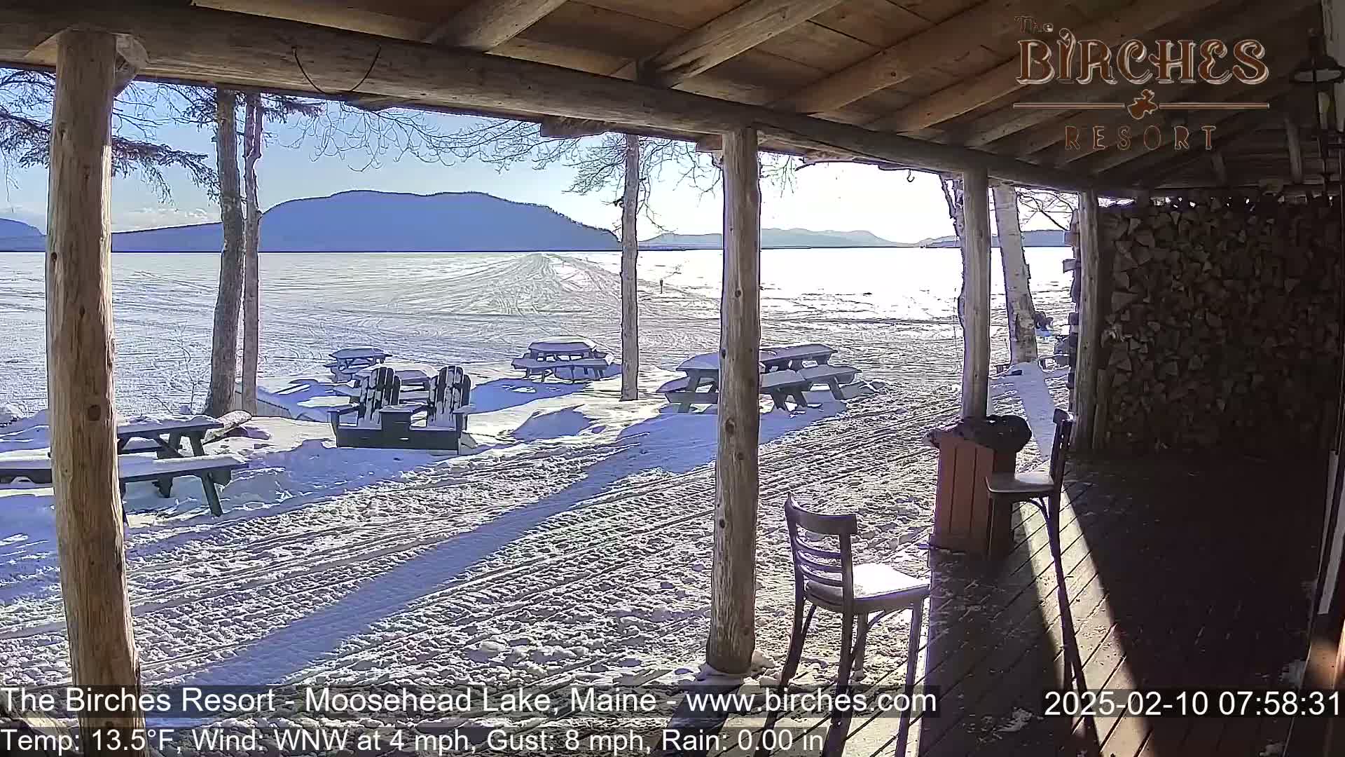 A sunny, snowy lakeside view is seen from a covered porch with picnic tables and chairs.