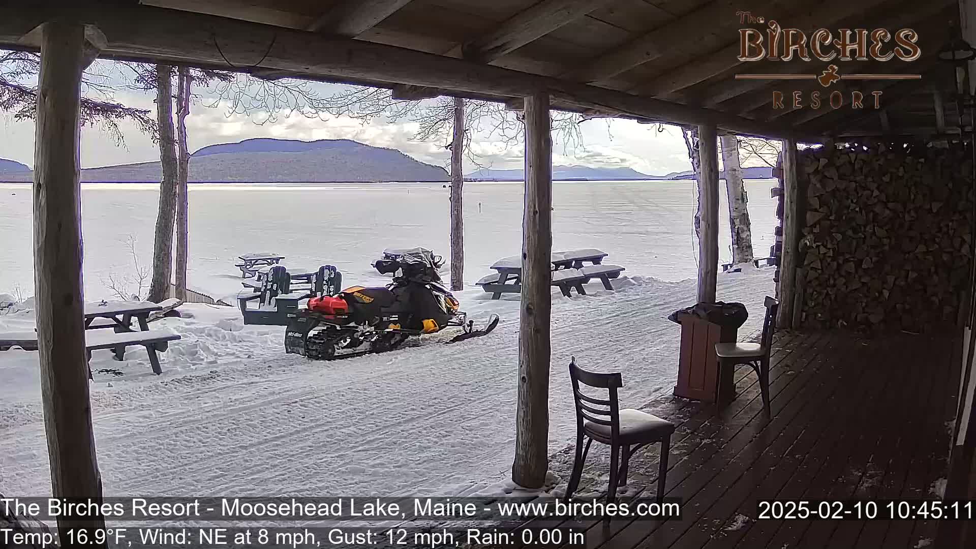 A snowmobile sits parked near snow-covered picnic tables on a porch overlooking a frozen lake and distant mountains under a partly cloudy sky.