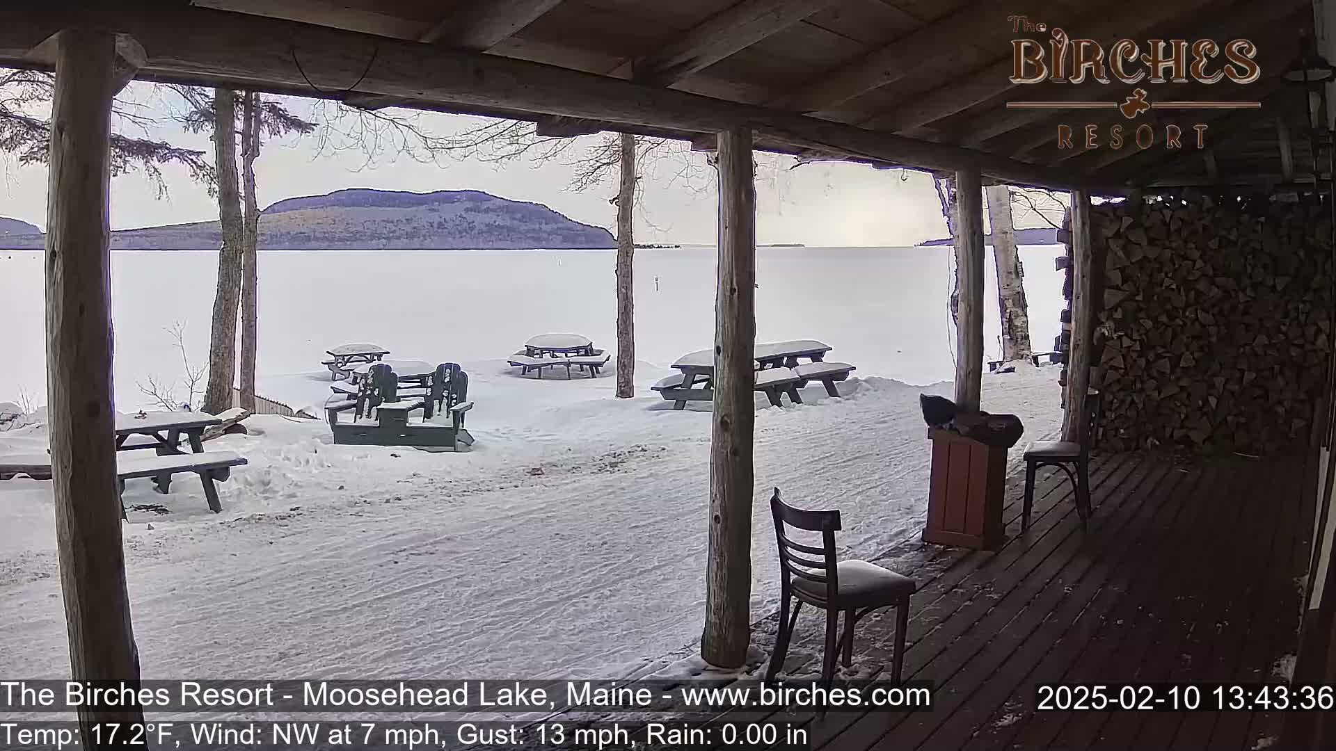 A snow-covered lakeside patio with several picnic tables and chairs is viewed from a porch, under a cloudy sky.