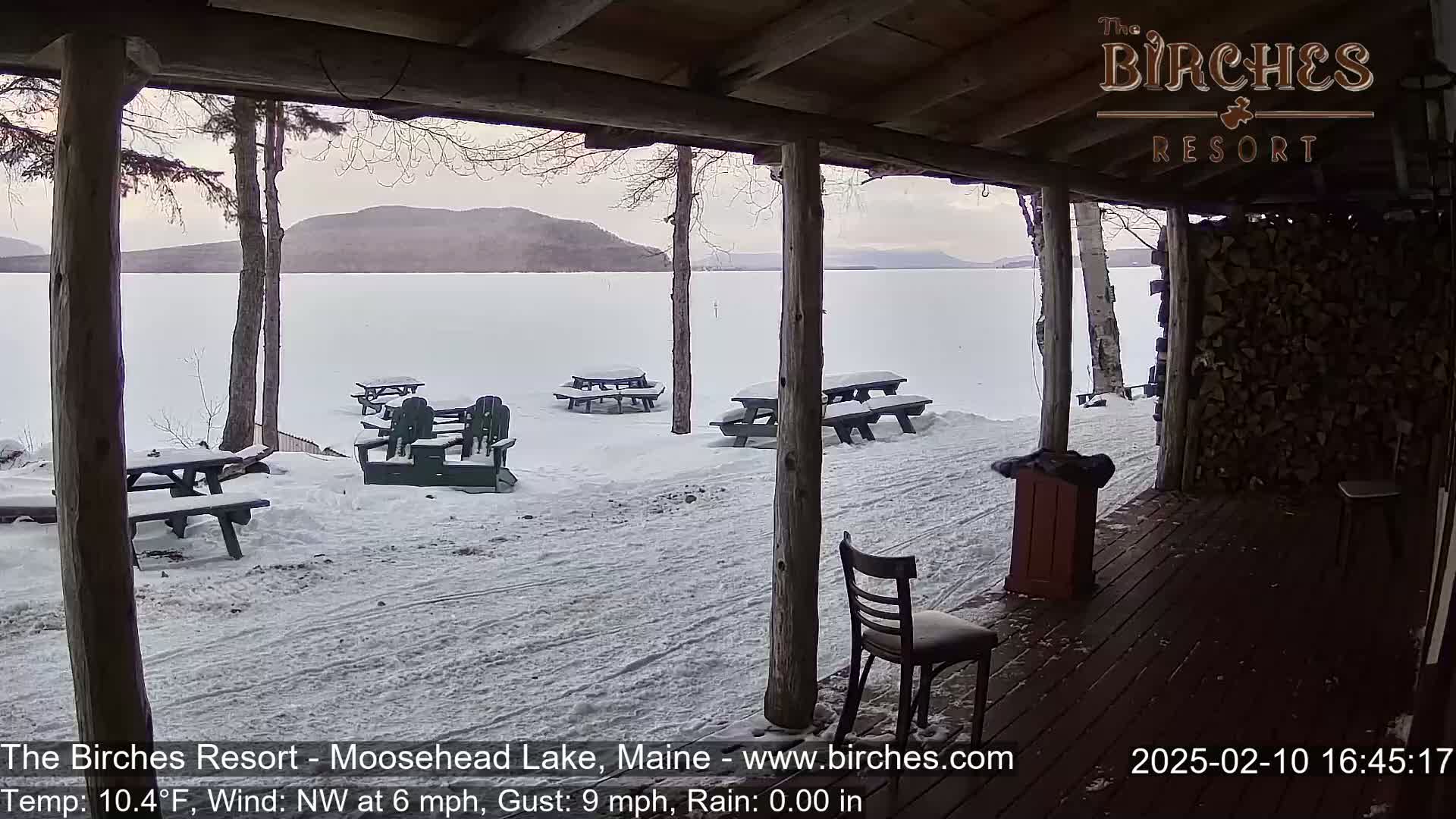 A snow-covered lakeside patio with picnic tables and chairs is viewed from a wooden porch, overlooking a frozen lake and distant mountains under a cloudy sky.
