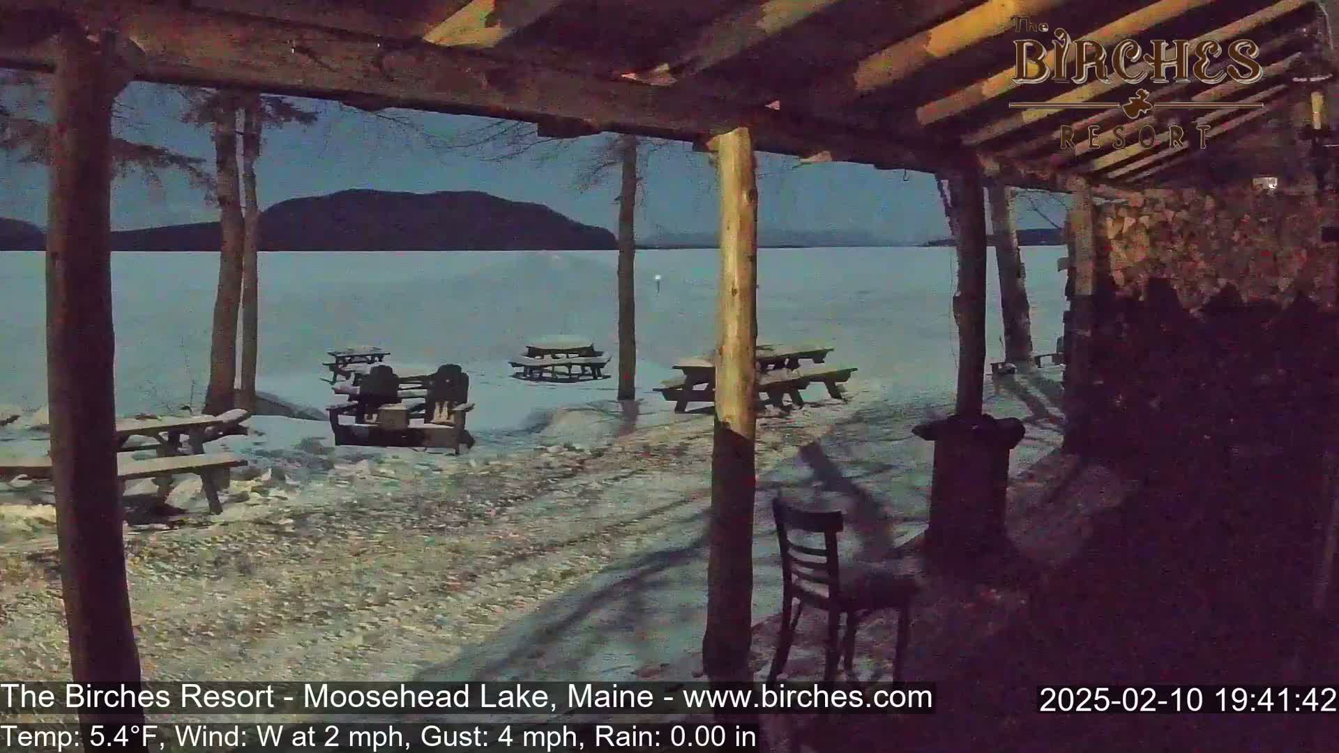 A snow-covered lakeside patio with picnic tables and chairs under a nighttime sky, viewed from under a wooden structure.