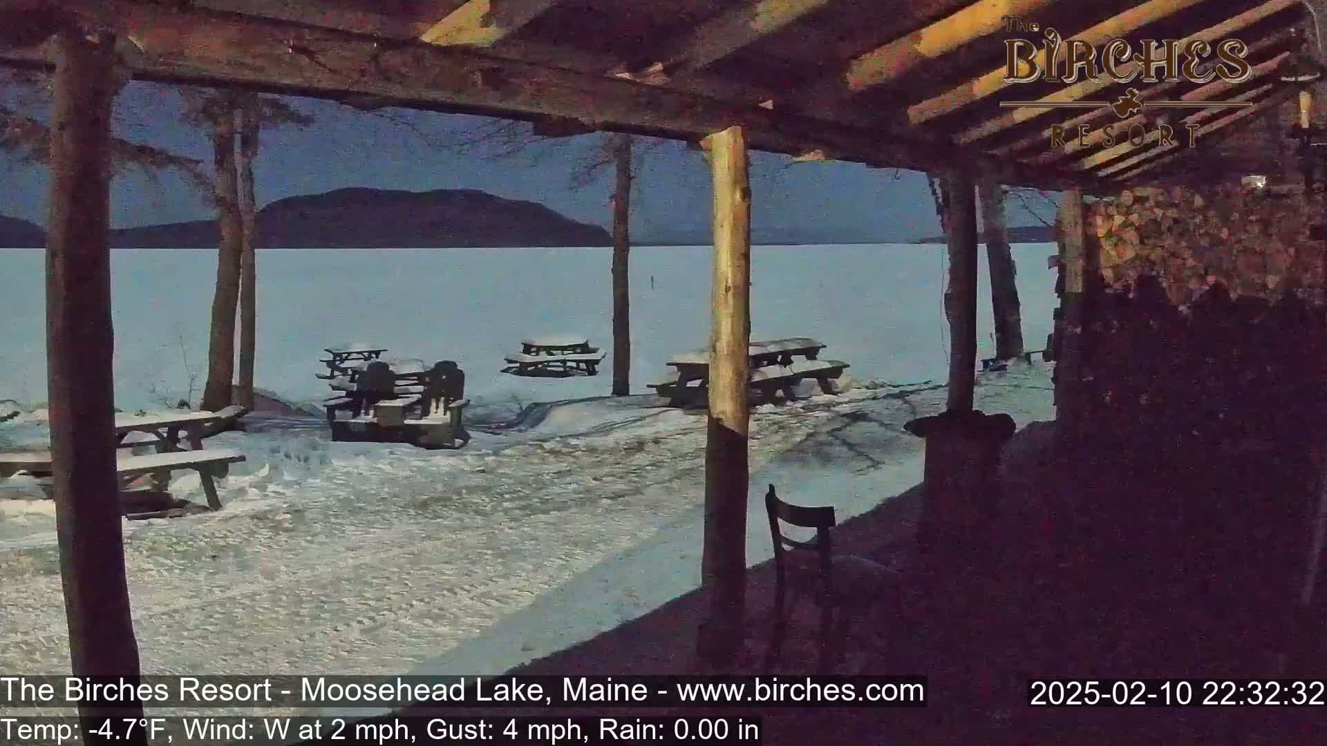 A snow-covered lakeside area is viewed from a covered porch at night, with several empty picnic tables and a chair visible under a dark sky.