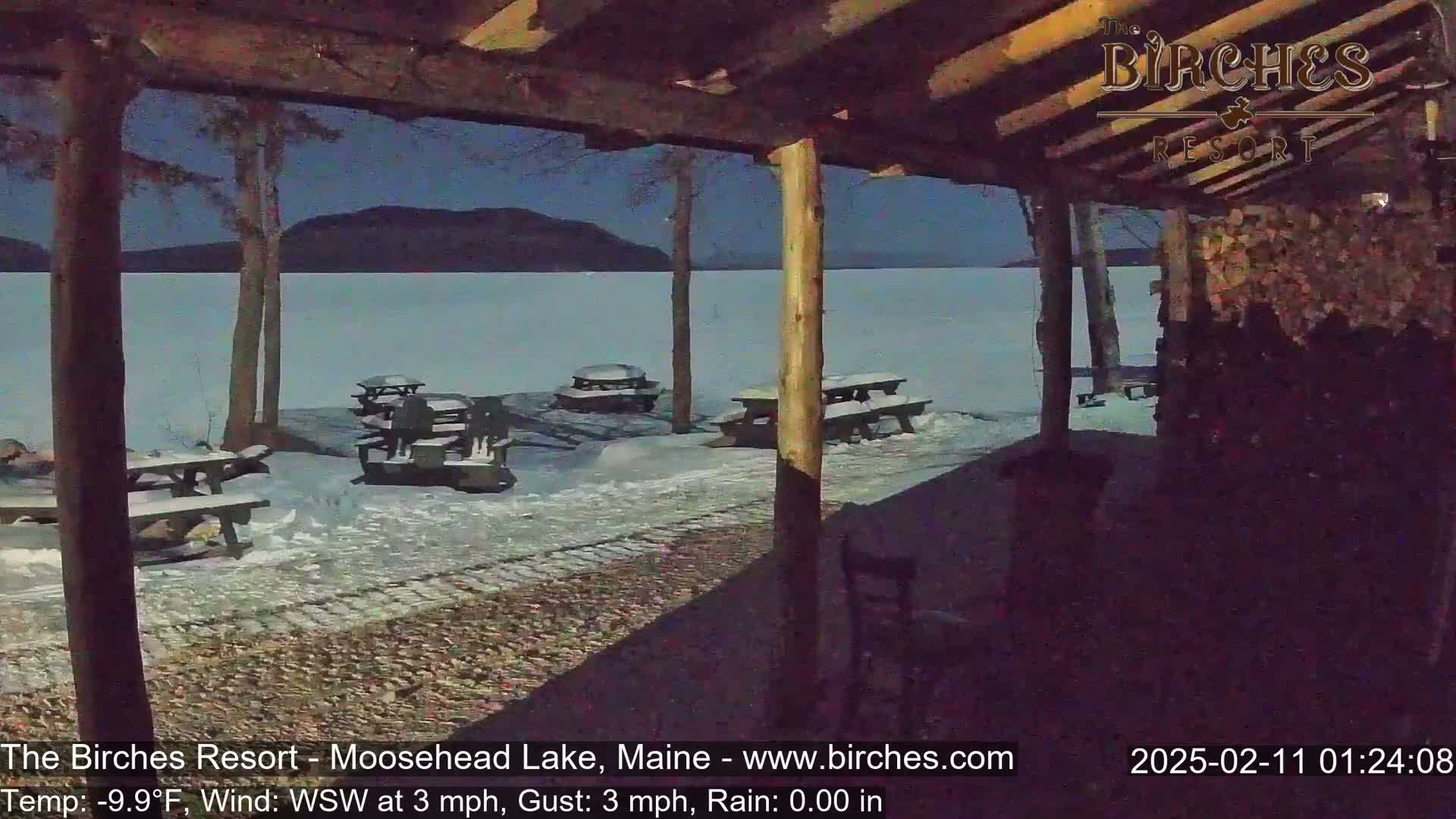 A snow-covered lakeside area with picnic tables is seen from under a wooden shelter at night.