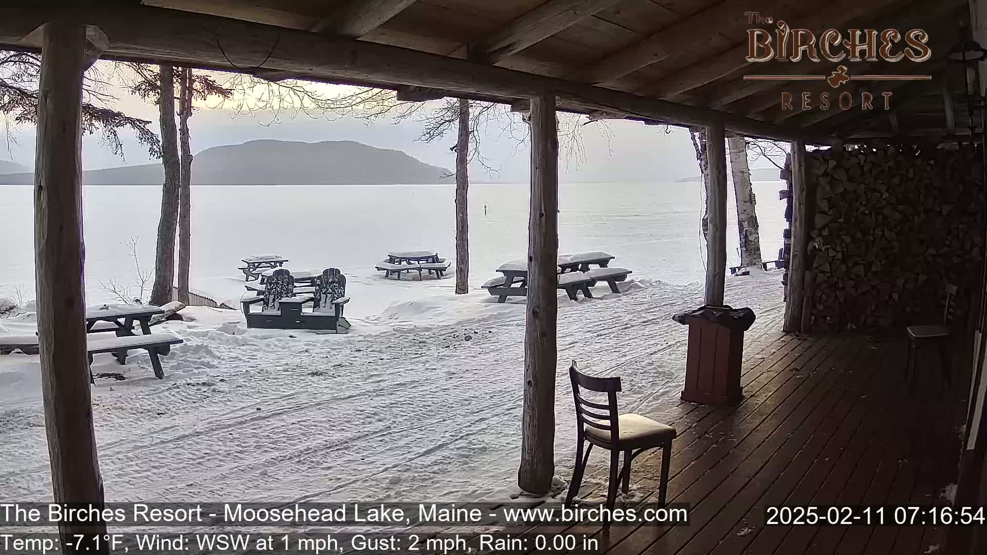 A snow-covered lakeside patio with picnic tables and Adirondack chairs is viewed from a covered porch on a cold, windy, and slightly rainy day.