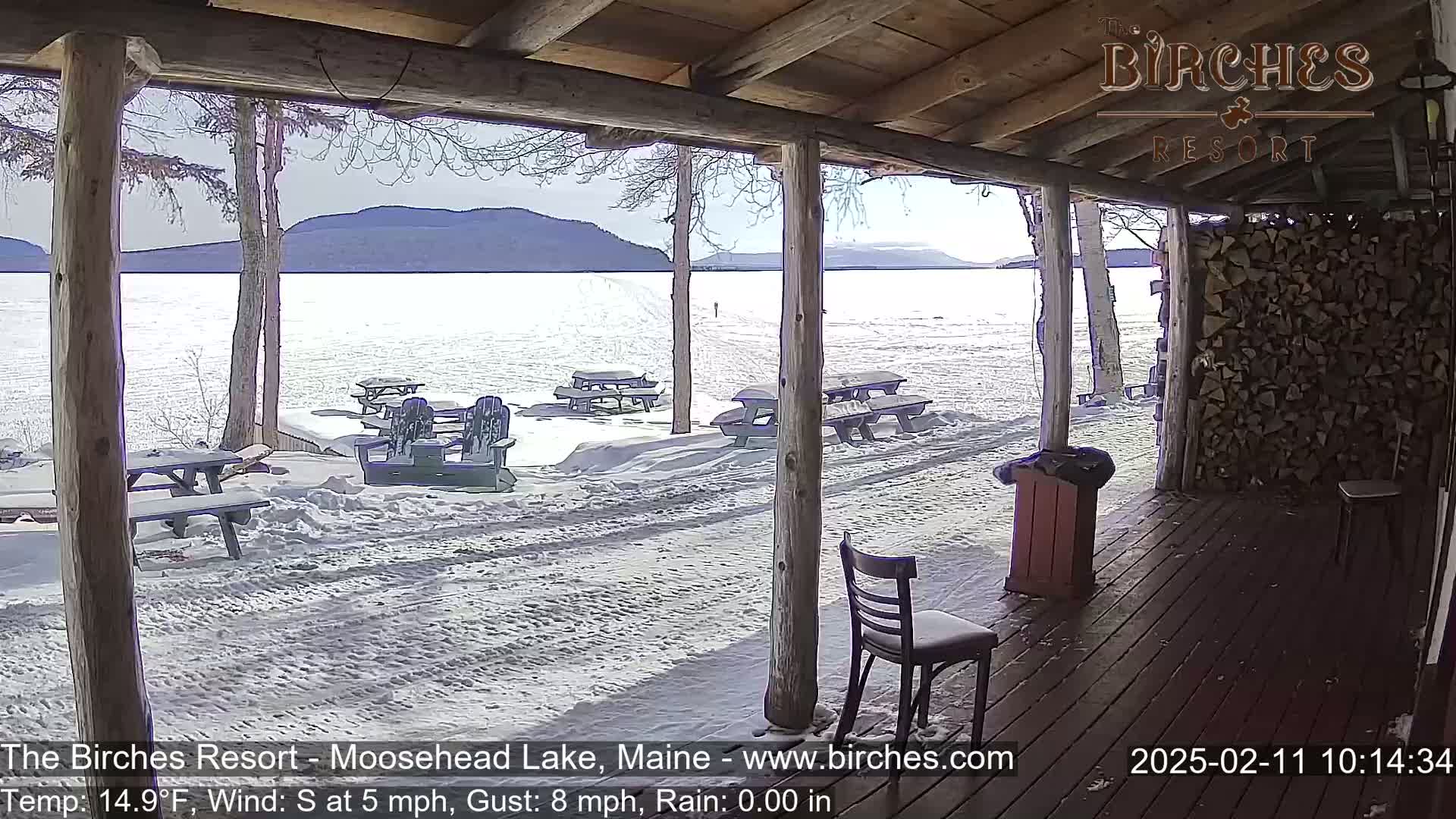 A snow-covered lakeside patio with picnic tables and chairs is seen from a covered porch on a sunny, cold day with a light south wind.