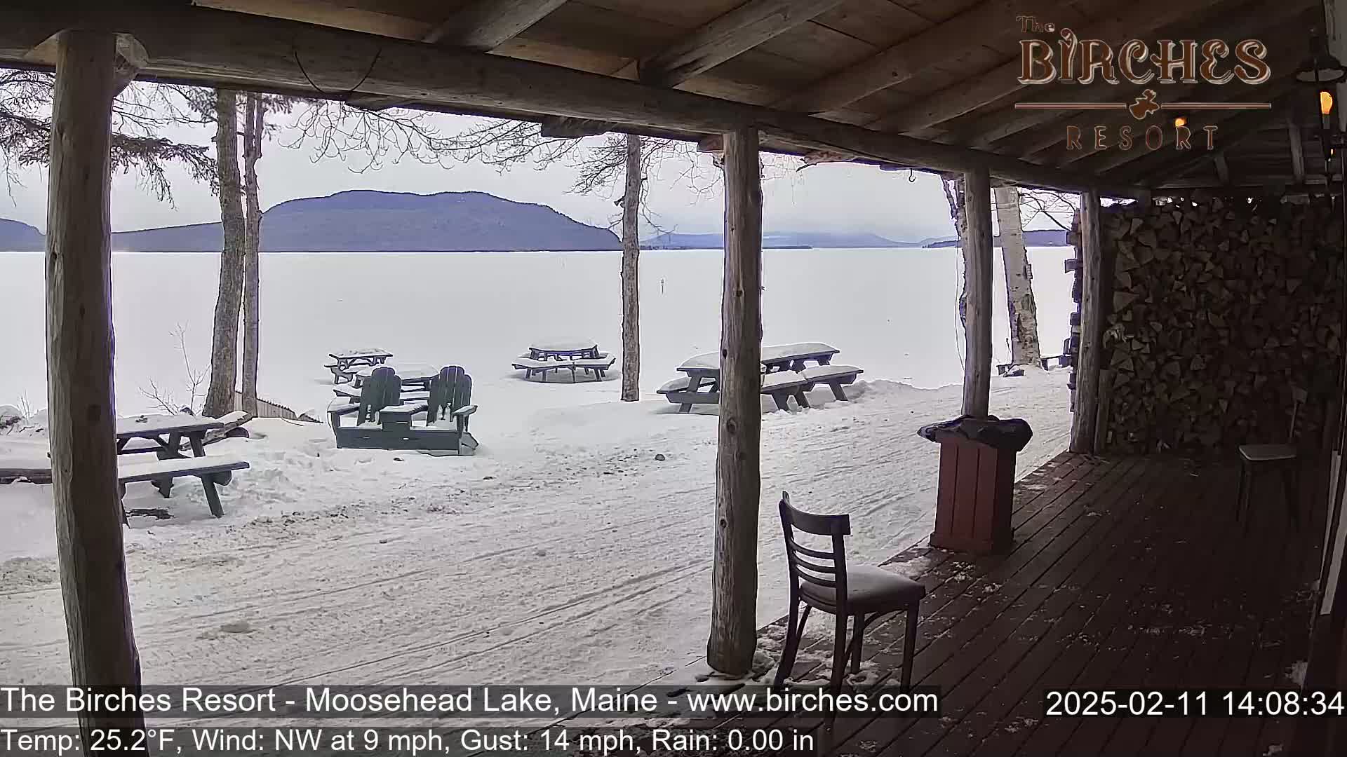 A snow-covered porch overlooks a frozen lake and distant mountains under a cloudy sky.