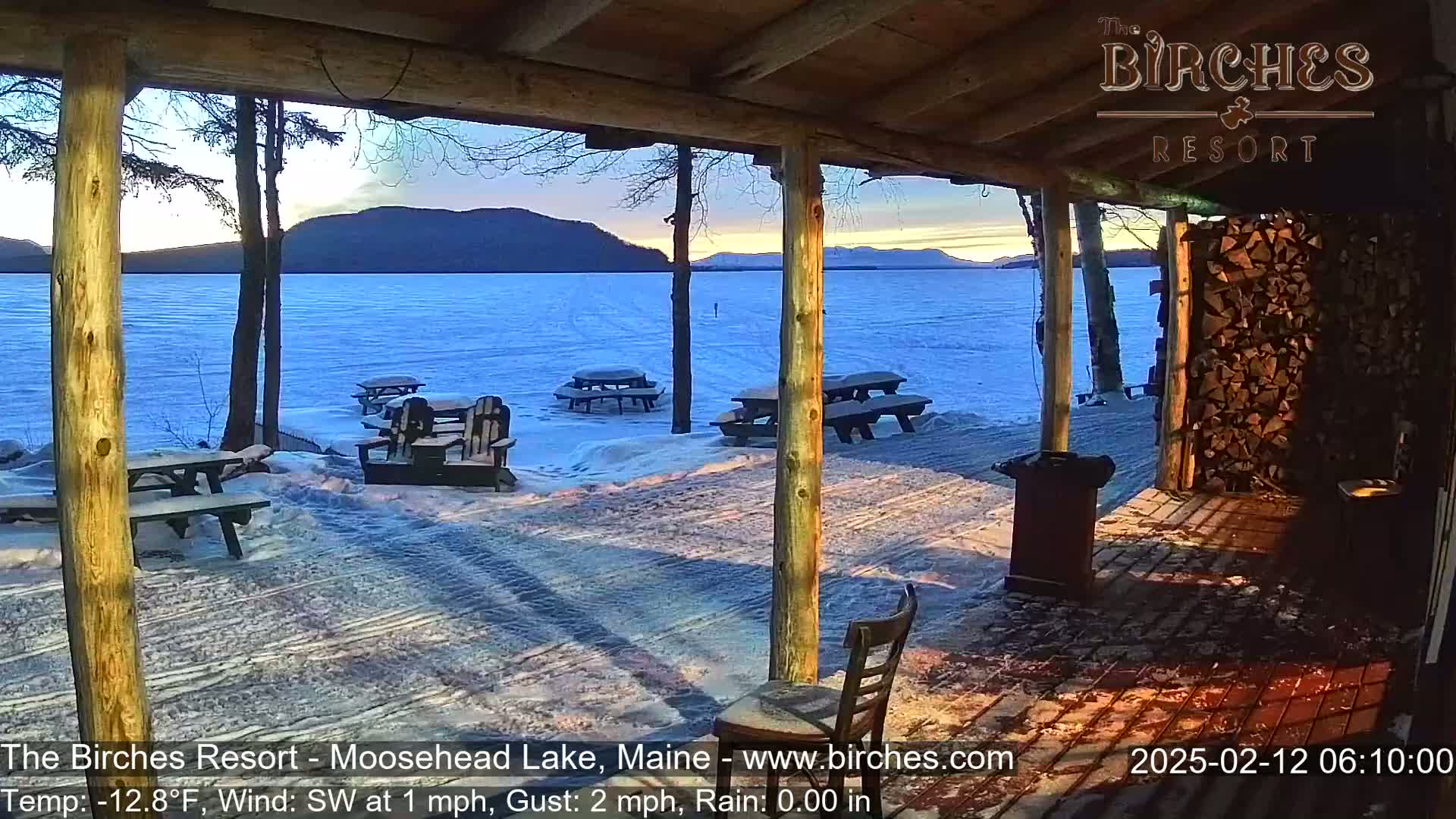A snow-covered lakeside patio with picnic tables and chairs, viewed from a covered porch, overlooks a frozen lake and mountains under a twilight sky.