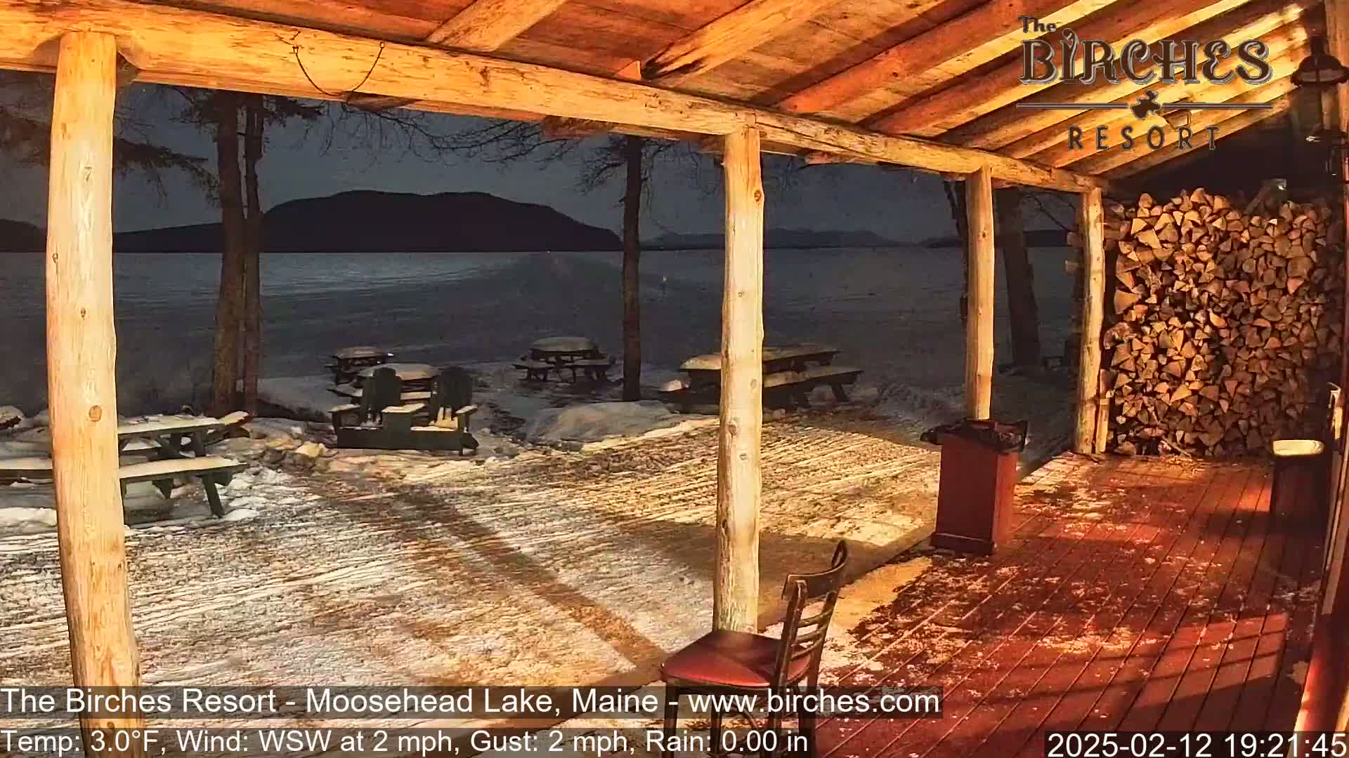 A snow-covered outdoor patio with picnic tables is seen from inside a wooden structure overlooking a calm lake under a dark night sky.