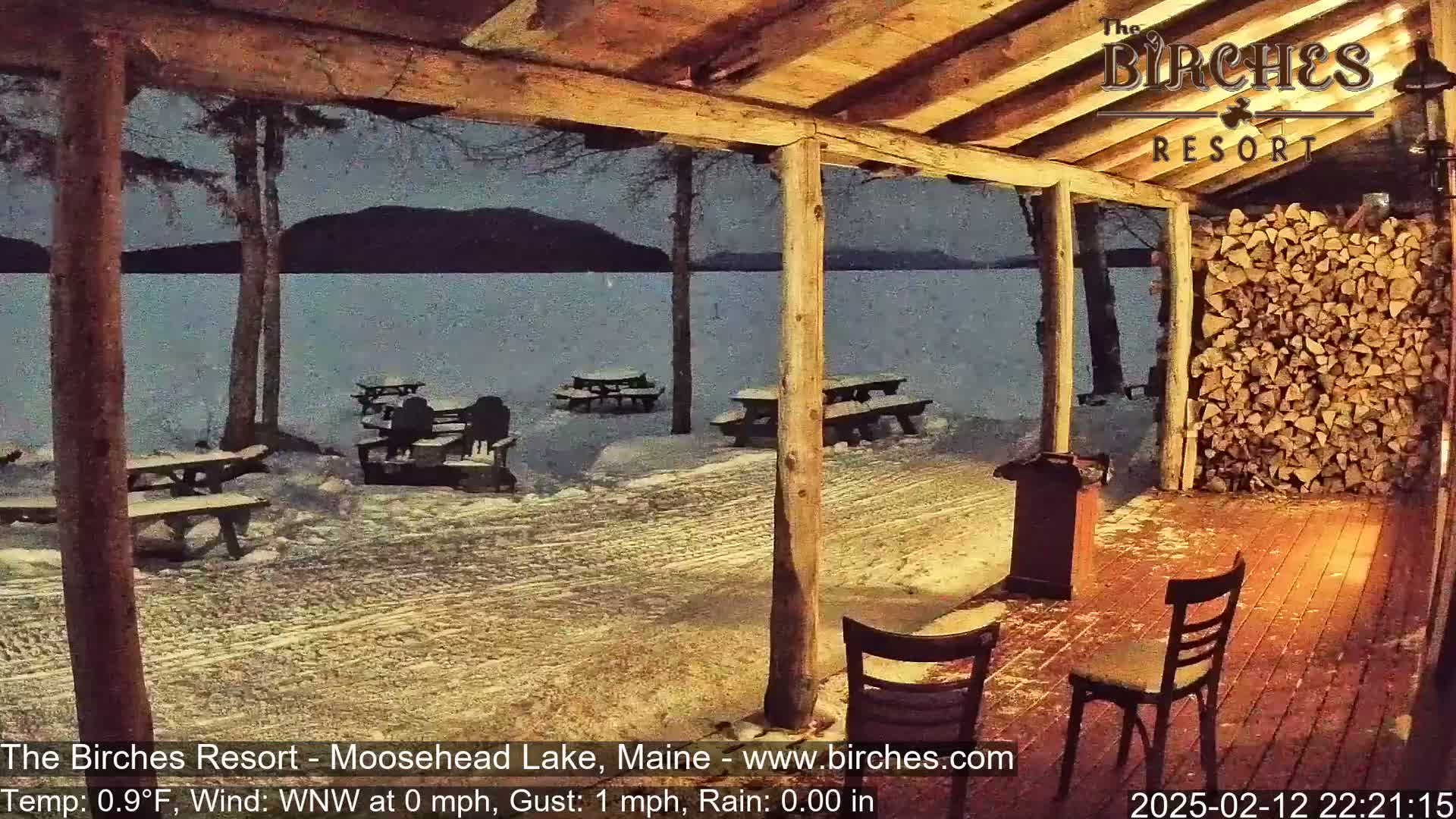 A snow-covered lakeside patio with picnic tables is viewed from a wooden porch at night, under light rain and a near-calm wind.