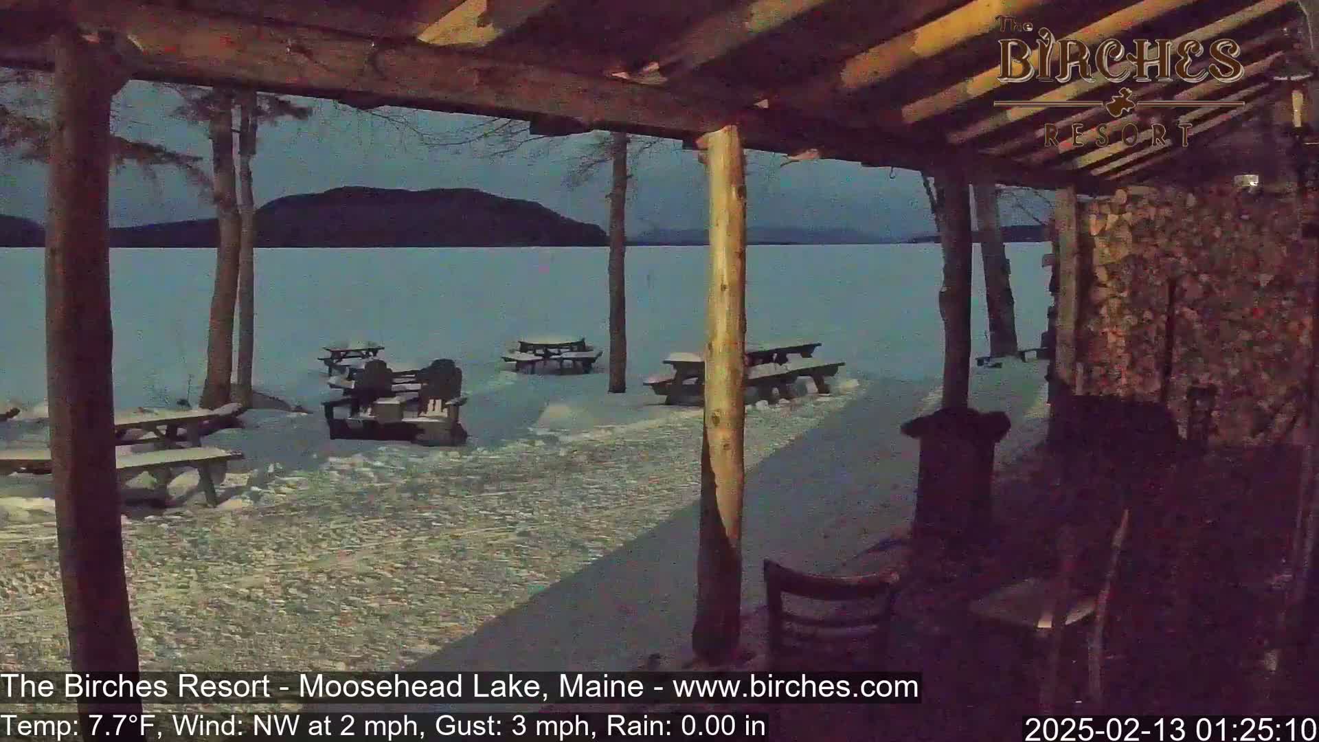 Under a covered porch, several picnic tables sit empty on a snow-covered lakeshore at night during a light rain.