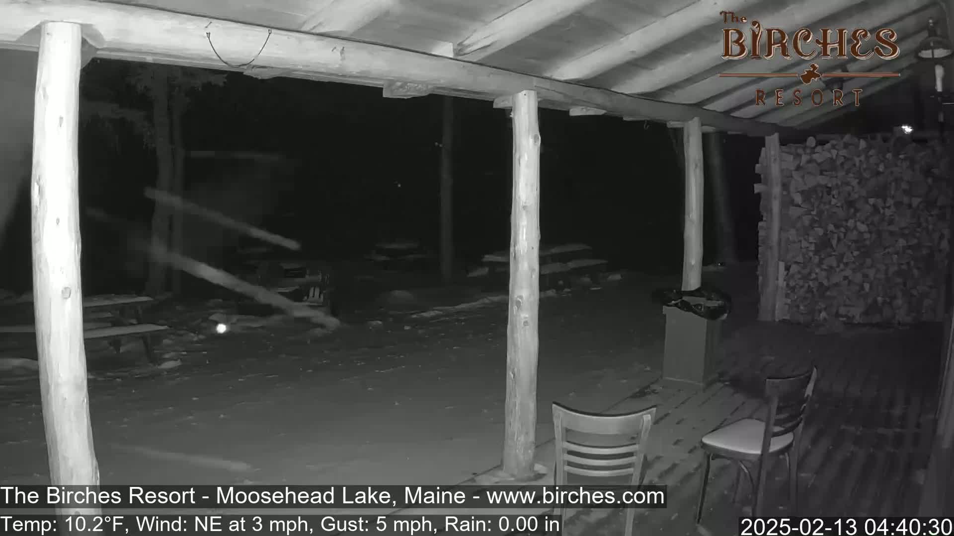 A nighttime, snowy scene shows an outdoor covered patio area with a few chairs and a woodpile, under a northeast wind with light gusts of snow.