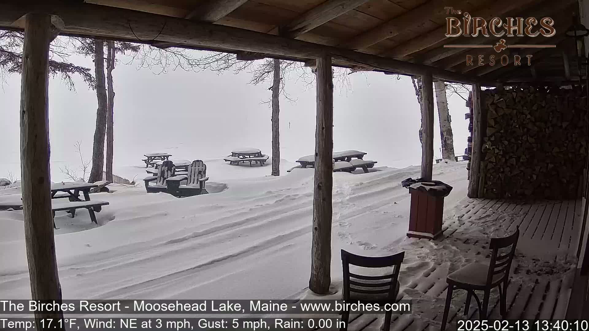 A snow-covered outdoor patio with picnic tables and chairs is viewed from a covered porch in overcast weather.