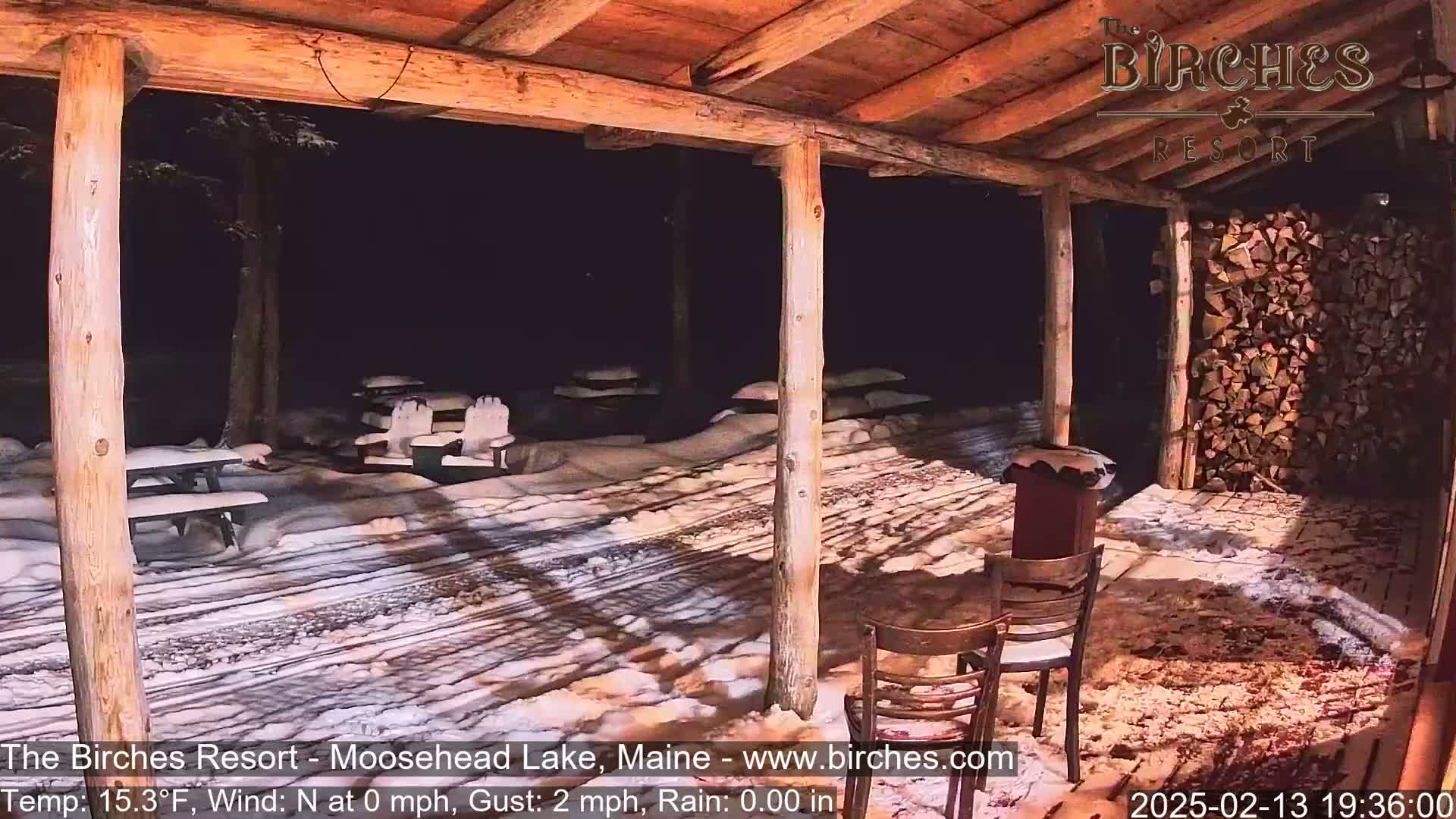 A snow-covered outdoor patio with wooden picnic tables and chairs is seen at night under a wooden shelter.
