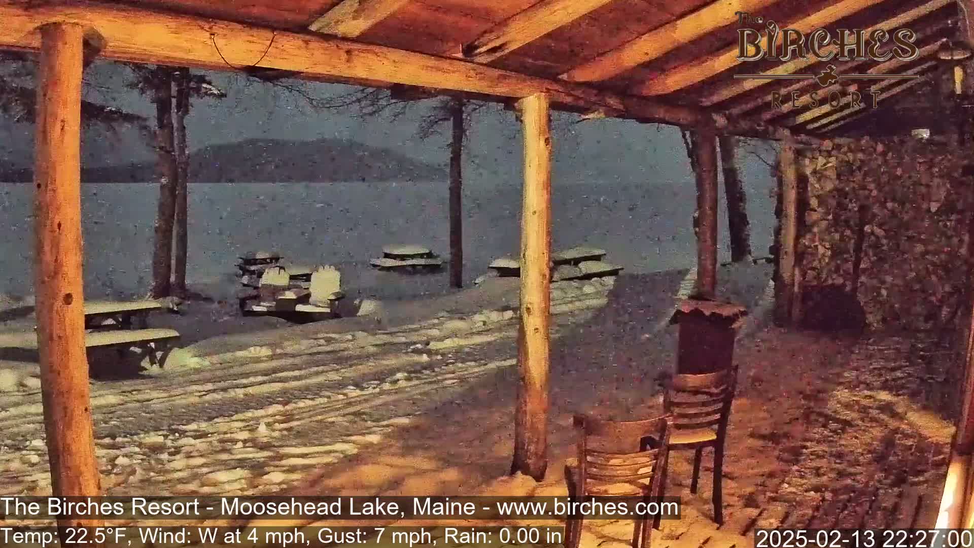 A snow-covered lakeside scene is viewed from a wooden porch at night, with picnic tables scattered across the snowy ground.