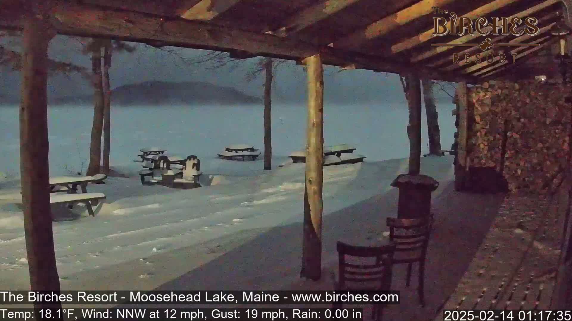 A snow-covered outdoor patio with picnic tables and chairs overlooks a lake under a twilight sky, with a northwest wind blowing at 12 mph and gusts up to 19 mph.