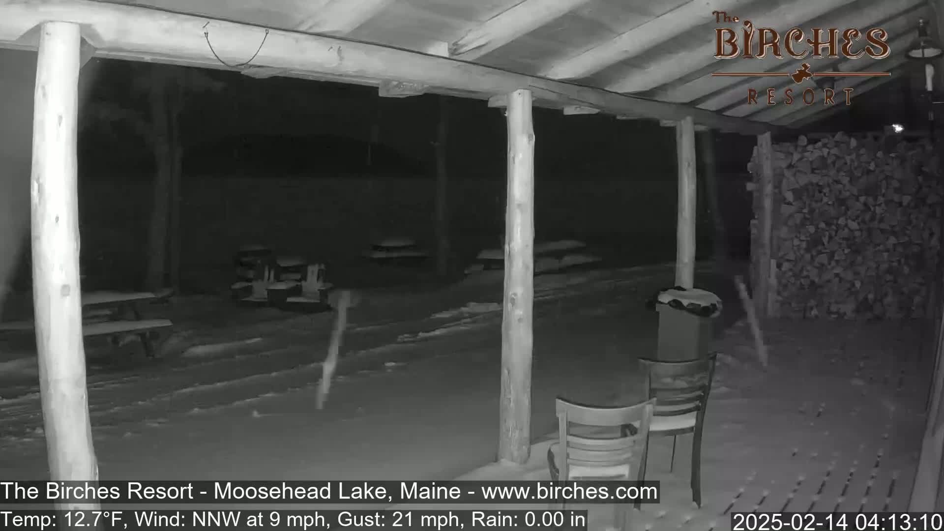 A snow-covered outdoor patio area at night, viewed from under a covered porch, with a strong wind blowing.