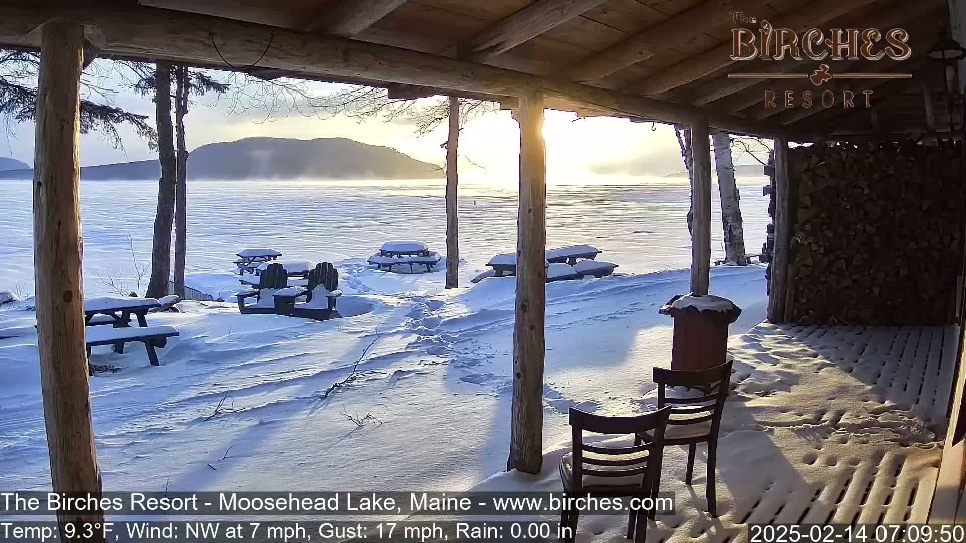 A snow-covered porch overlooks a frozen lake with mountains in the distance under a partly sunny sky.