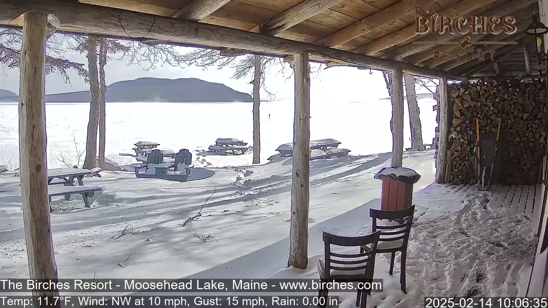 A snow-covered porch overlooks a frozen lake with mountains in the distance under a sunny sky.