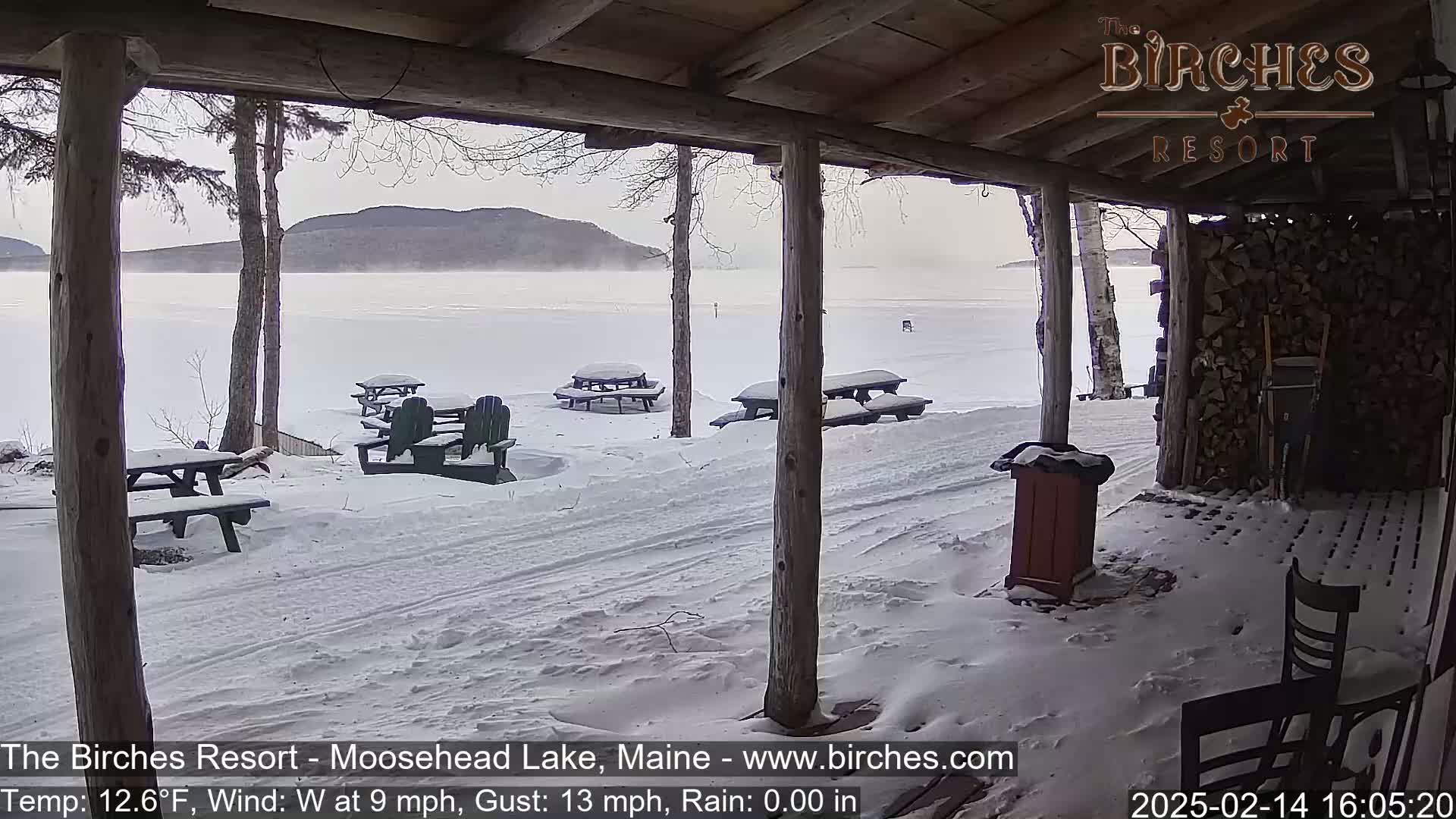 A snow-covered porch overlooks a frozen lake with distant mountains under a hazy sky, with several picnic tables and chairs covered in snow.