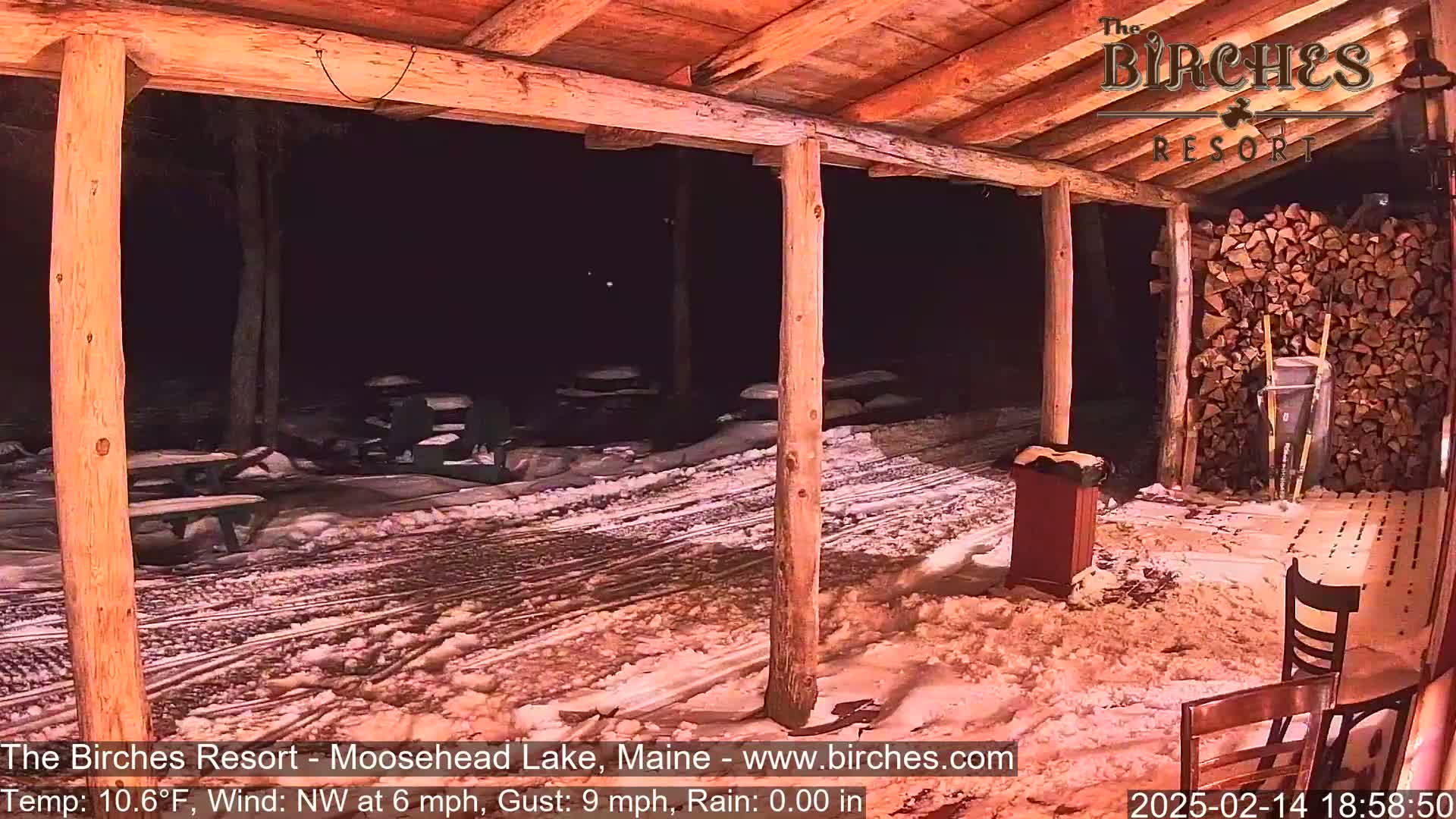 A snow-covered outdoor patio area at night, viewed from inside a rustic wooden structure, with picnic tables and a woodpile visible.