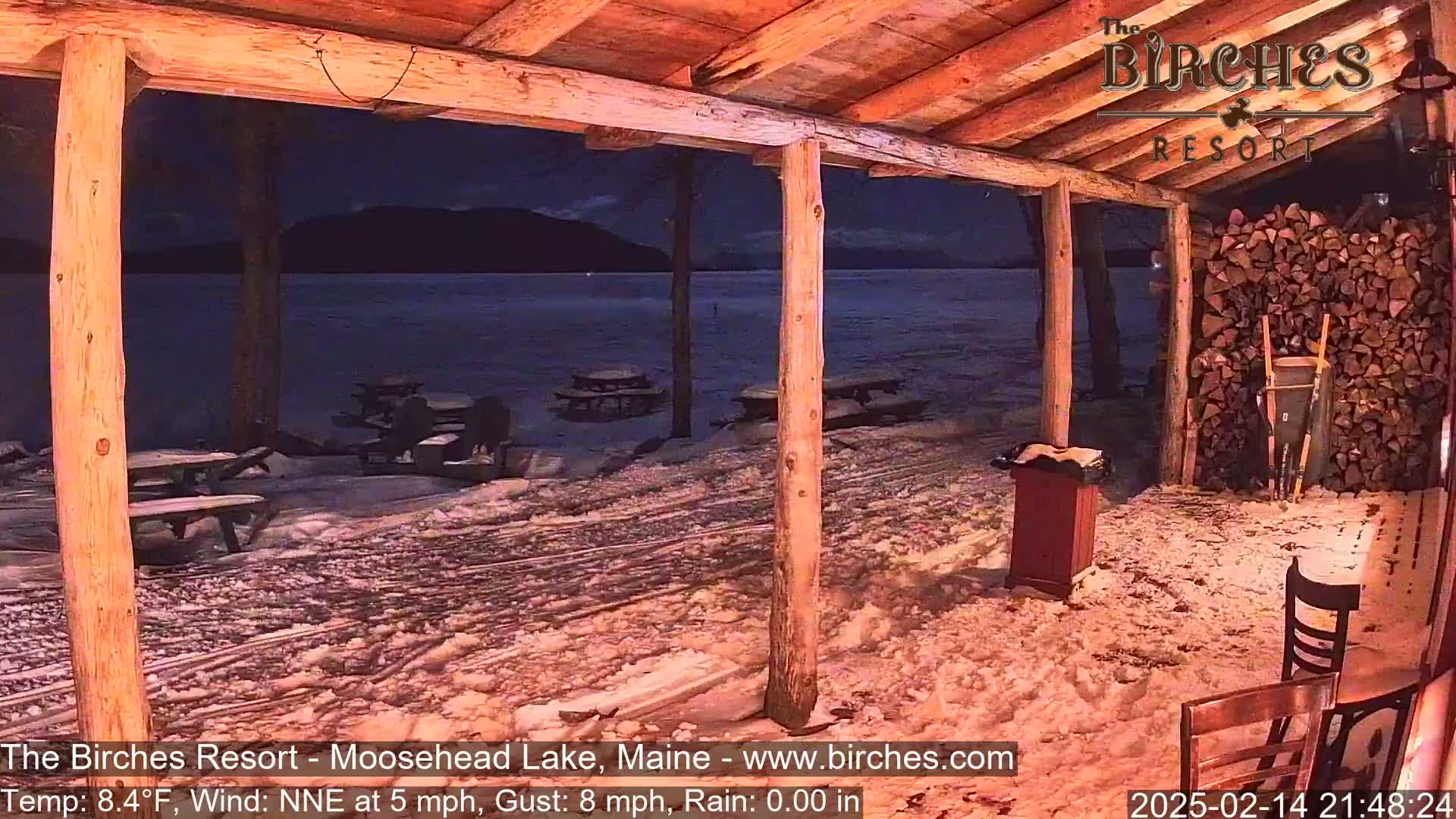 A snow-covered outdoor patio with picnic tables overlooks a dark lake under a night sky.