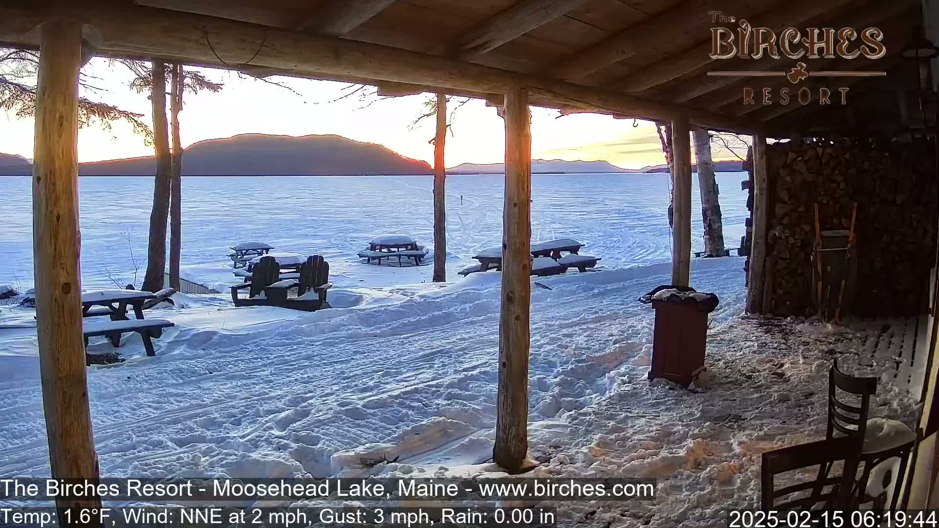 A snow-covered lakeside porch with picnic tables and a view of a frozen lake and mountains at sunset.
