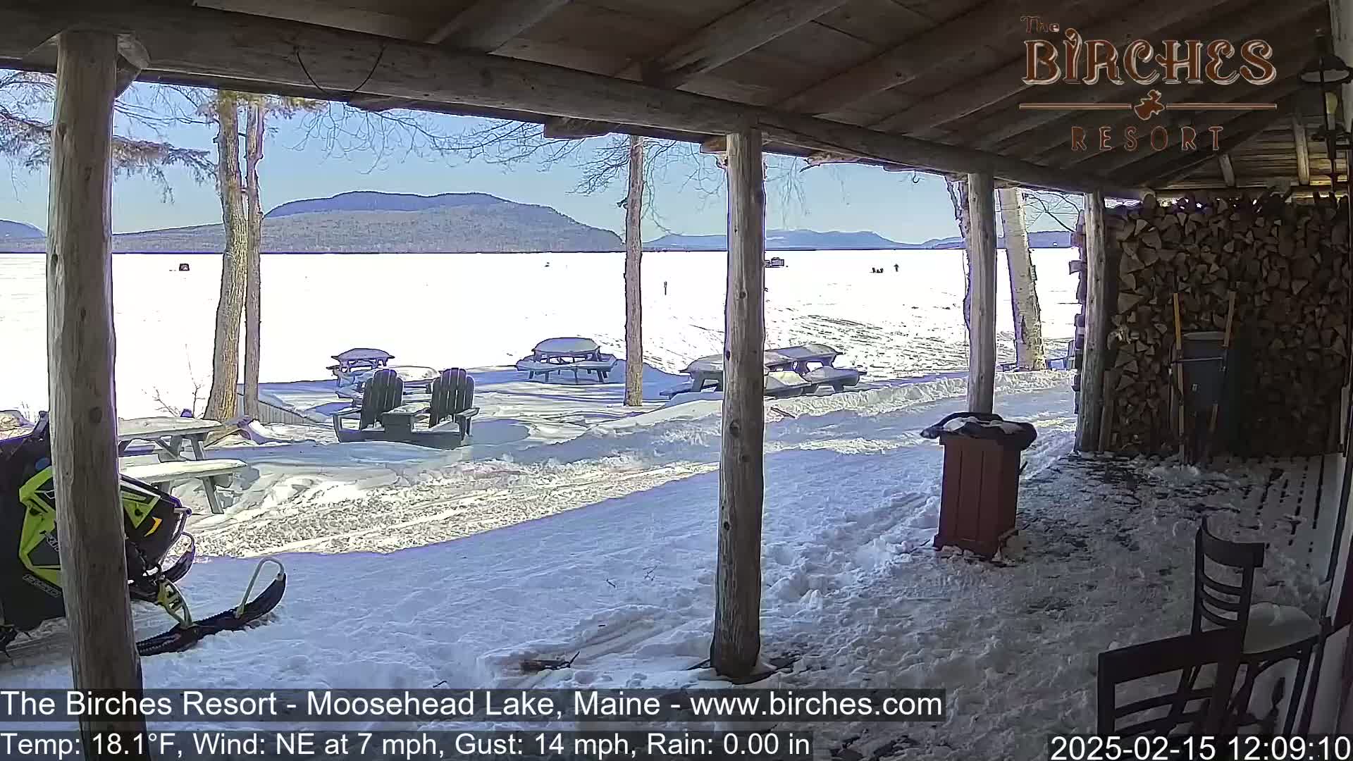 A snow-covered lakeside patio with picnic tables and a snowmobile is viewed from a covered porch on a sunny day with a northeast wind.
