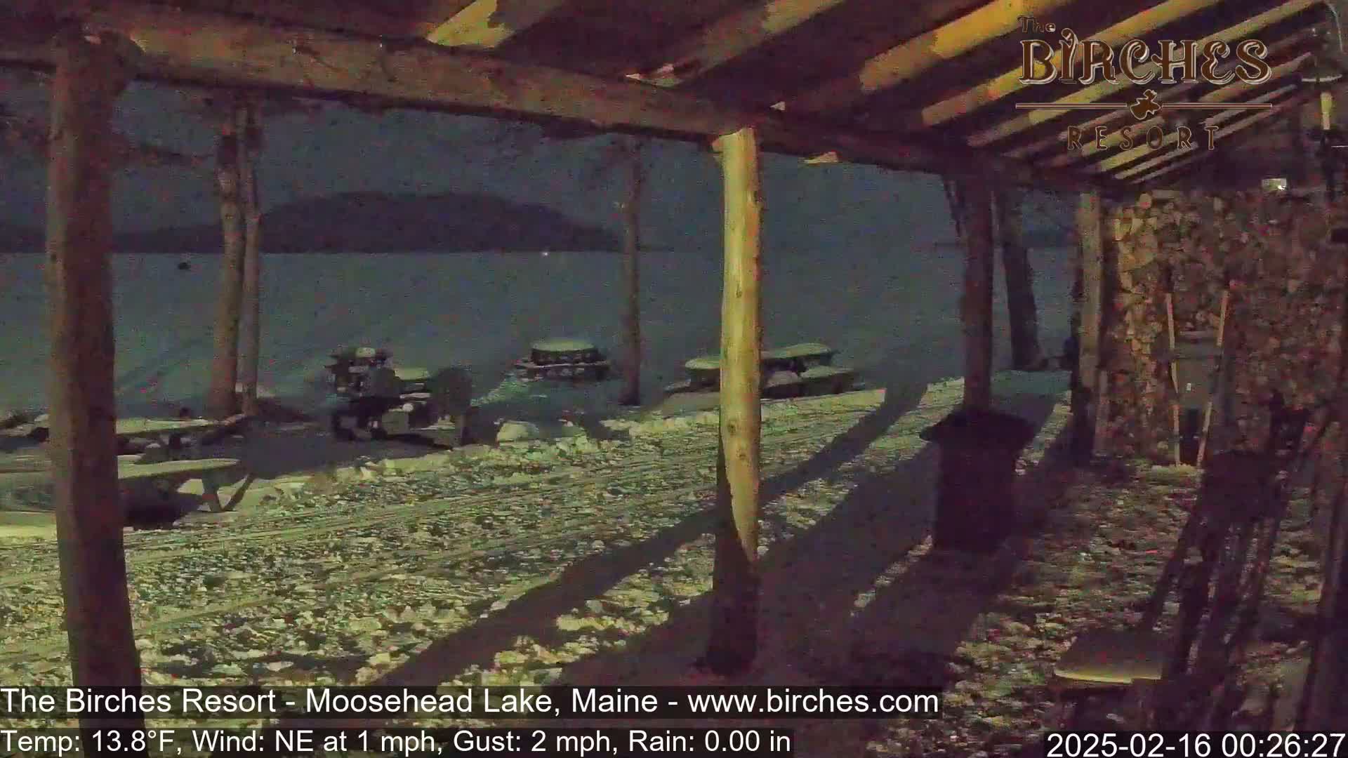 A snow-covered outdoor area with picnic tables is viewed from under a wooden structure at night, during a light rain.