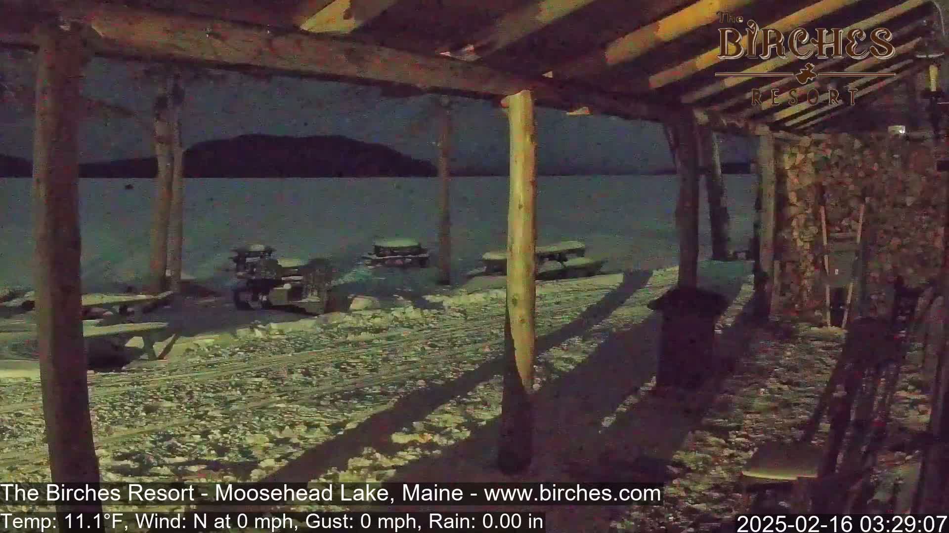 A snow-covered lakeside area is viewed from under a wooden structure at night, with several picnic tables visible.