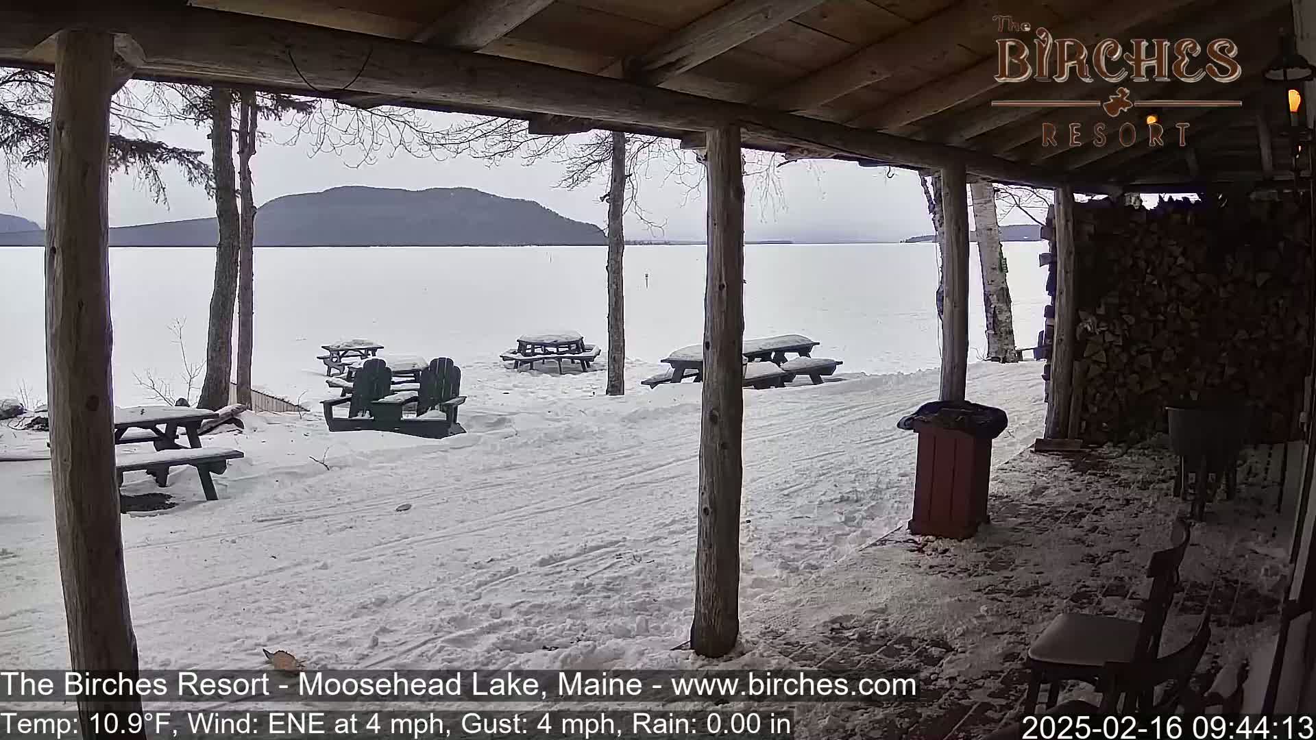 Snow-covered picnic tables sit outside a covered porch overlooking a snow-covered lake and distant mountains under an overcast sky.